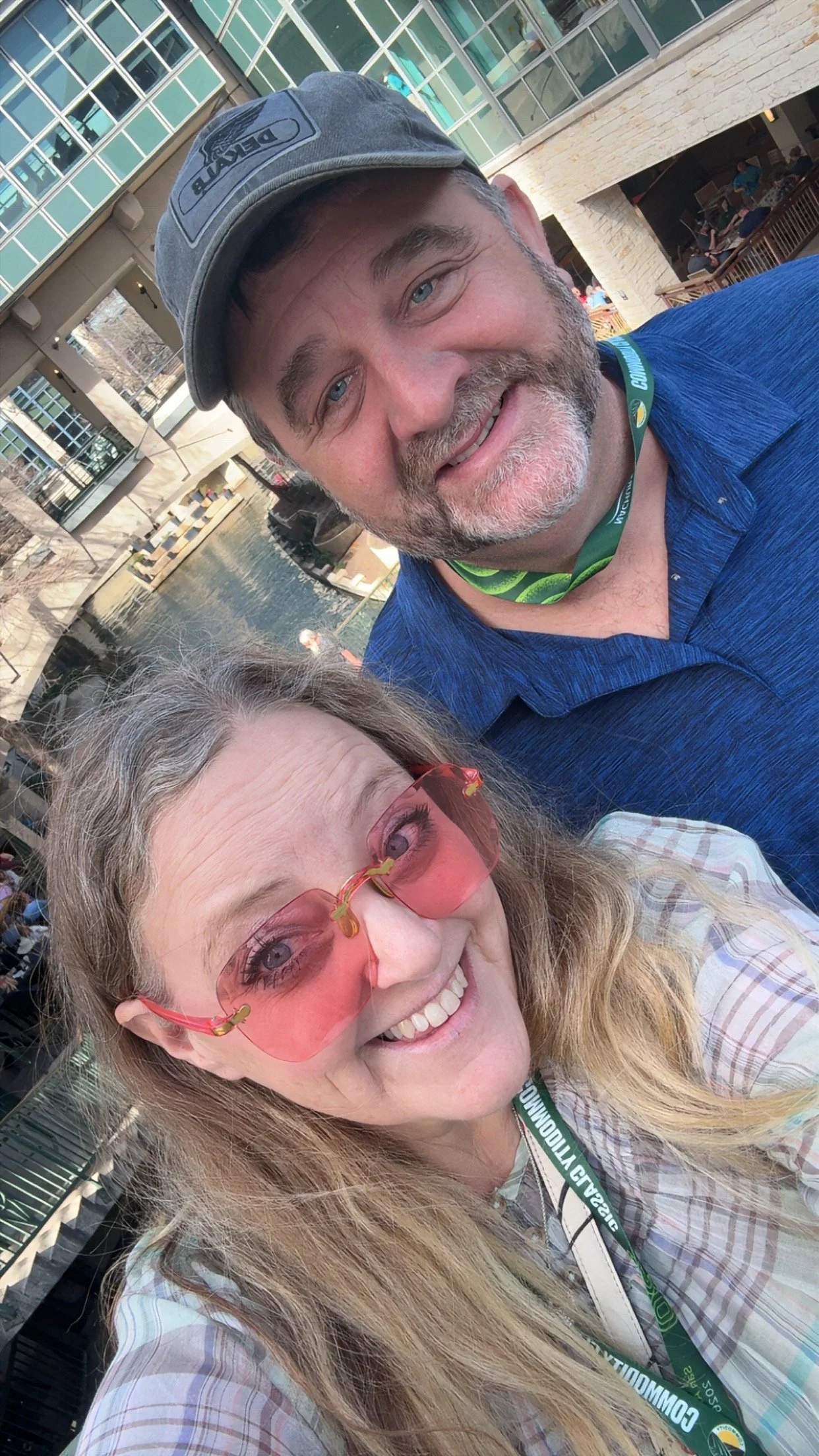 A selfie of a man and a woman outdoors near a water feature with a modern building in the background. The man wears a gray cap and blue shirt, the woman wears pink sunglasses and a plaid shirt.