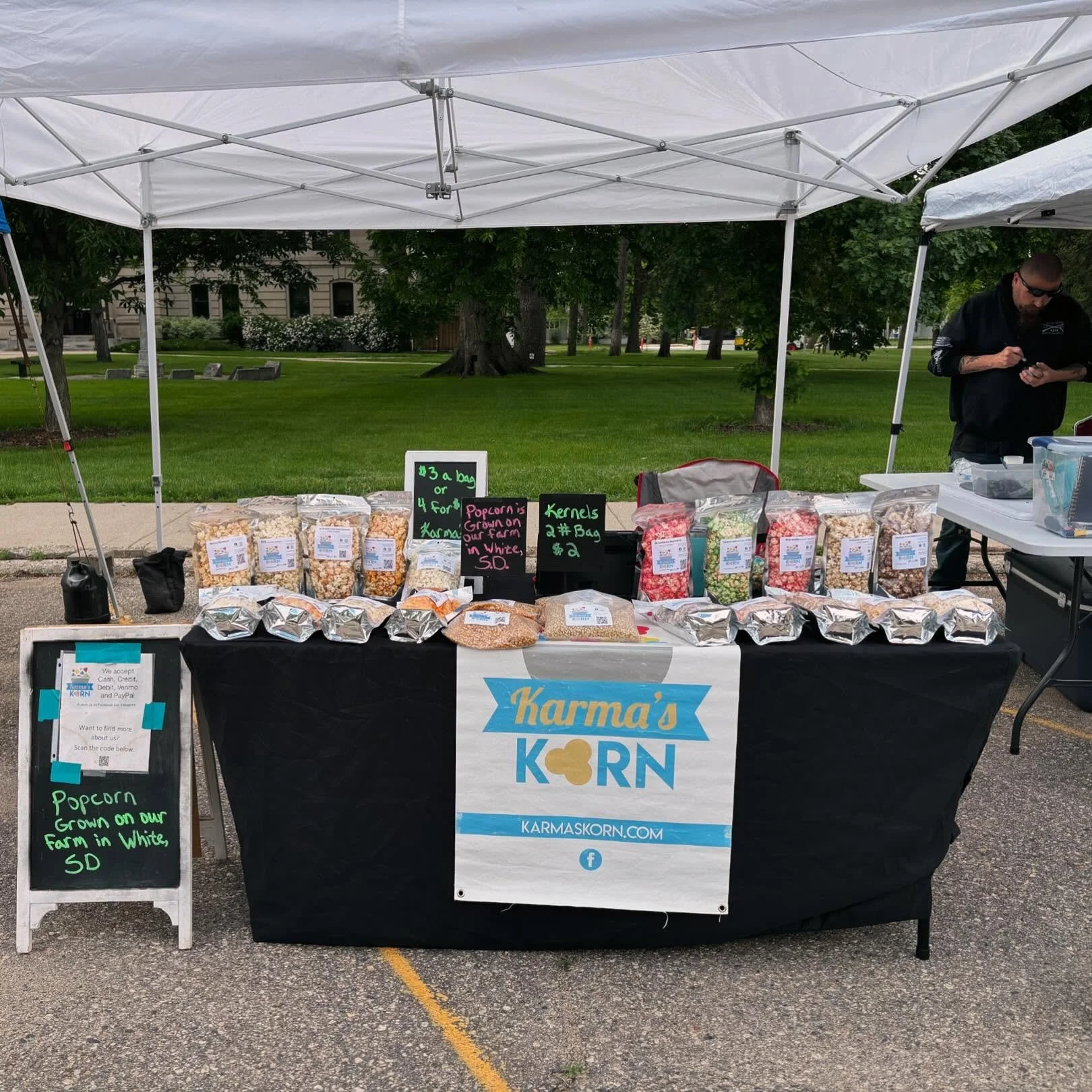 A booth at an outdoor market with a black tablecloth bearing a Karma's KORN logo. The table displays various bags of popcorn, some in clear plastic bags and others in foil. There's a chalkboard sign with green and pink writing noting prices and that the popcorn is grown on a farm in White, South Dakota. To the side, a man in dark clothing and sunglasses appears to be looking at his phone. A white canopy tent covers the booth, set against a background of trees and a grassy park area.