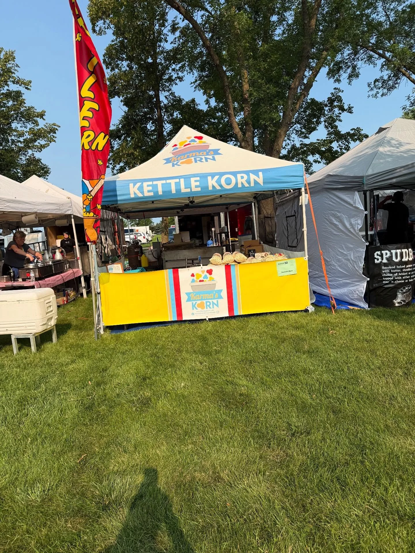 A tent at an outdoor festival with the name 'Karma's Korn' and a colorful banner. There are other tents and people in the background on a grassy field with trees and a blue sky.