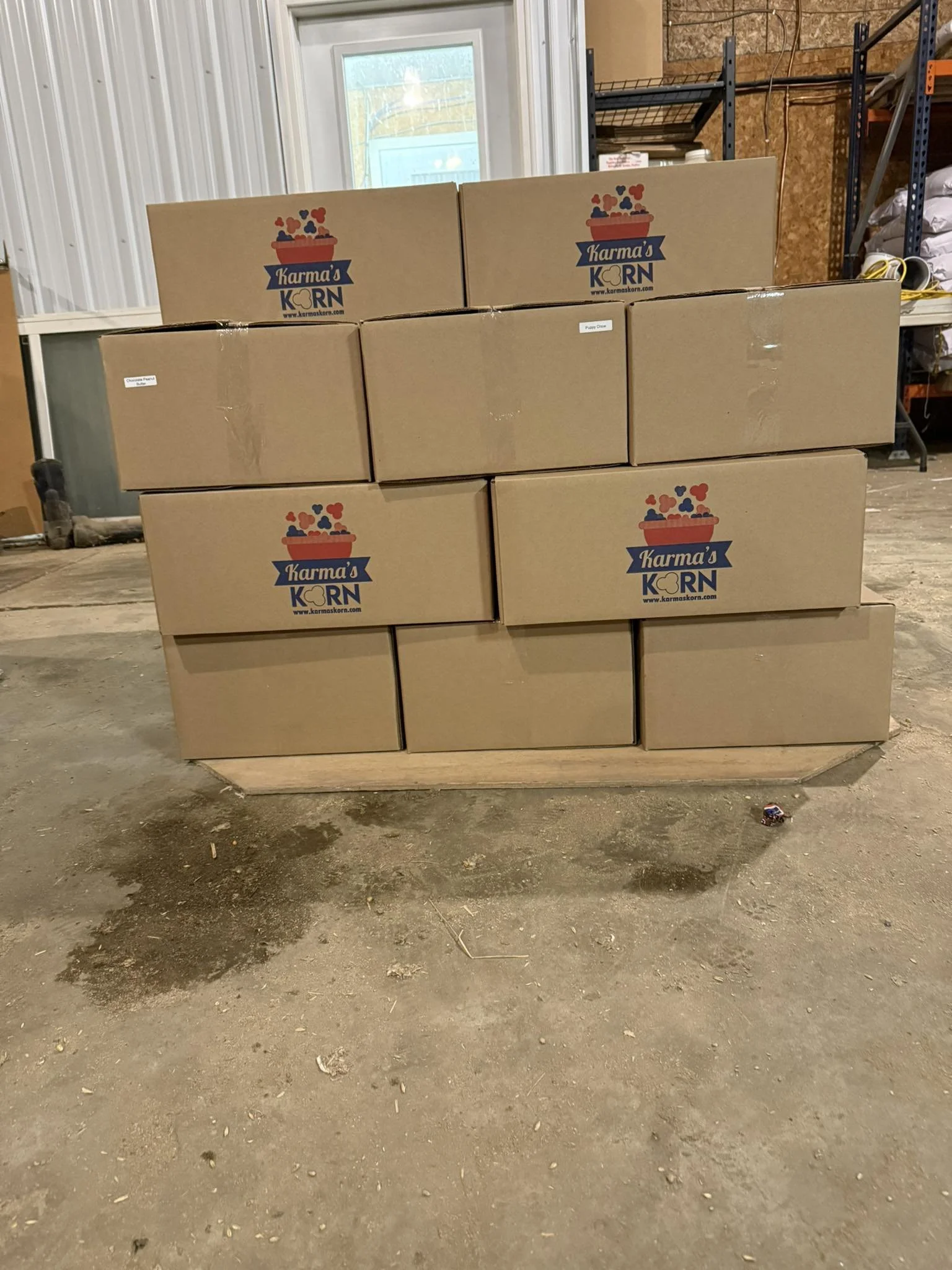 Stacked cardboard boxes with printed logo labeled 'Karma's KORN,' placed on a wooden pallet inside a warehouse.