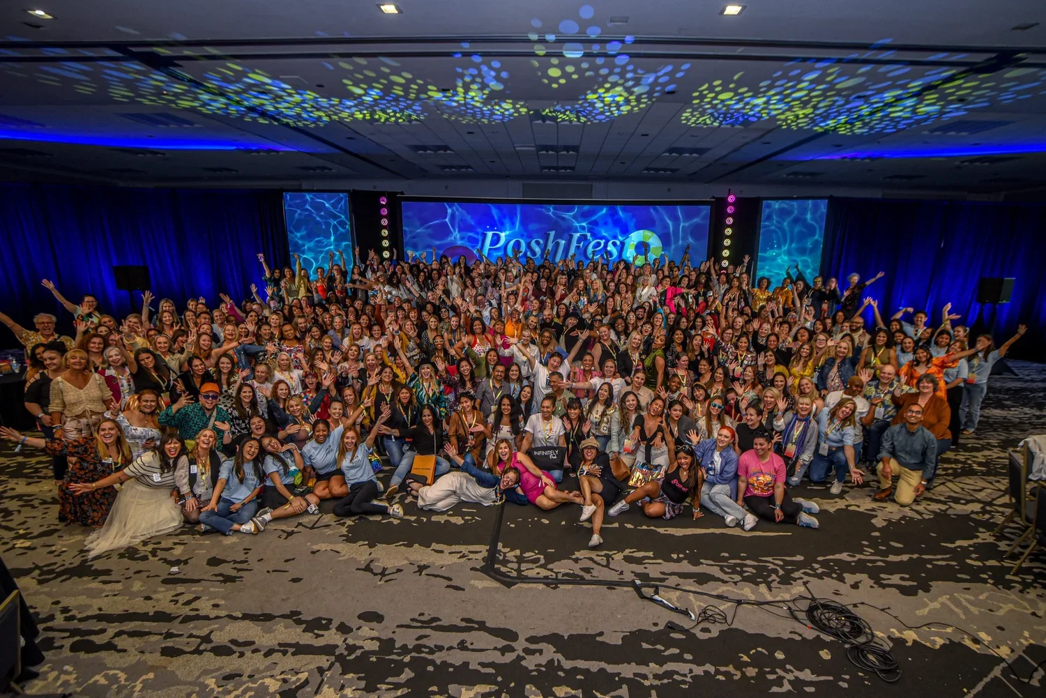 A large group of people on a stage, smiling and posing for a photo at a conference called Posh Fest. The background displays a digital screen with the event name, and the stage is decorated with blue lighting and curtains. The group includes people of diverse ages, ethnicities, and fashion styles, some sitting on the floor and others standing, with many raising their hands in celebration.