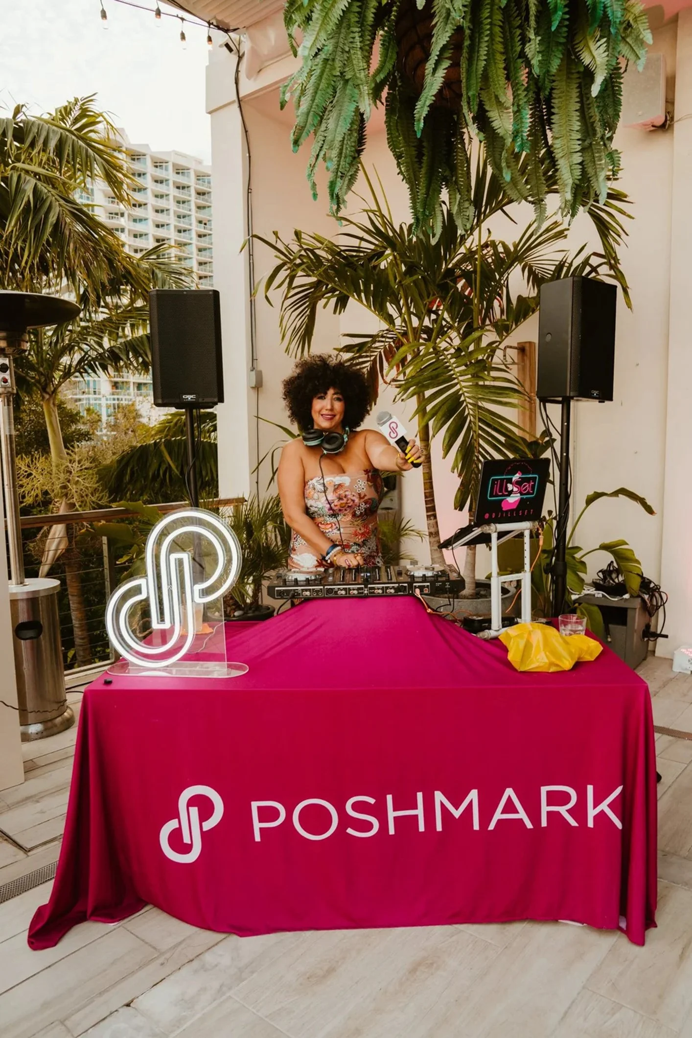 A woman with curly hair standing behind DJ equipment at an outdoor event. She is holding a microphone and wearing headphones around her neck. There is a pink tablecloth with the Poshmark logo on a table in front of her.
