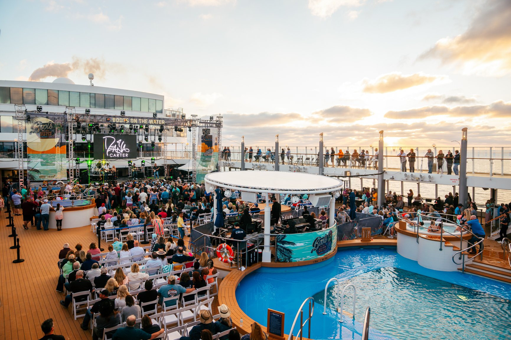 Crowd gathered on a cruise ship deck watching a live band perform on stage at sunset, with the ocean in the background.