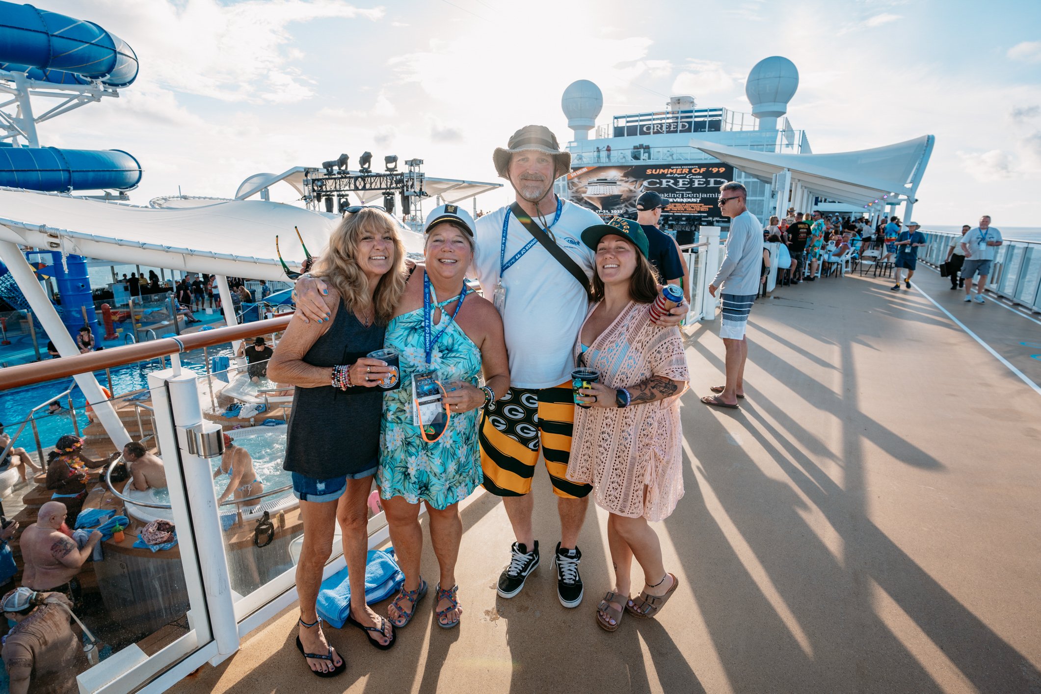 A group of four smiling people, three women and one man, standing together on the deck of a cruise ship. They are enjoying a sunny day. The background shows the cruise ship's water slide, pool area, and informational screens, with other cruise passen