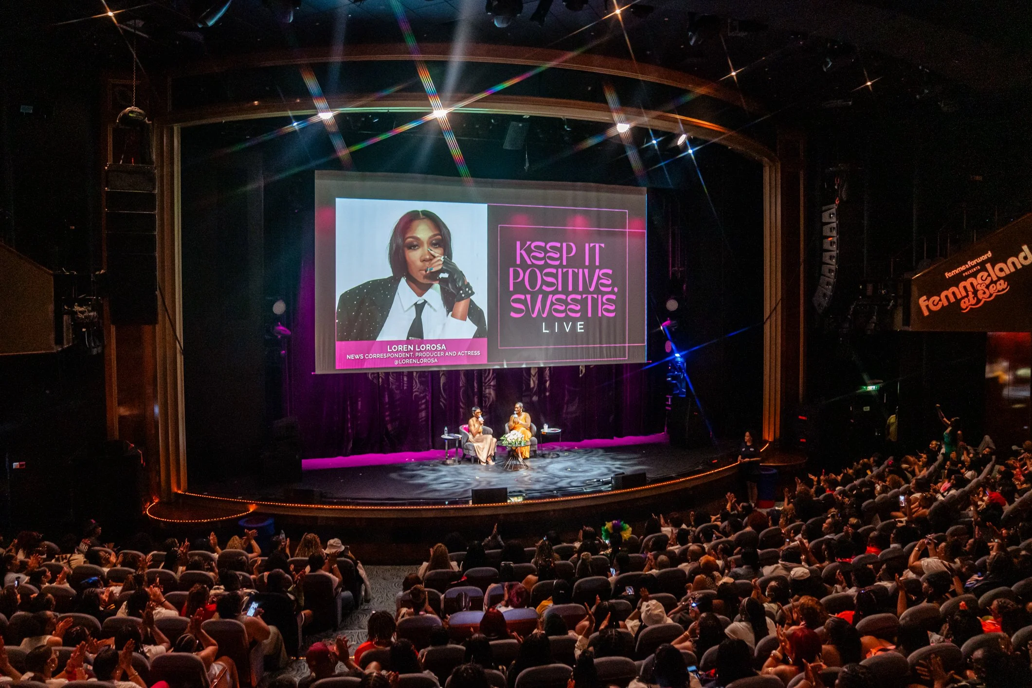 A stage at the Femmeland of Sea event with a large screen displaying a woman's portrait and text that reads "KSSP IT POSITIVES SWESTIS LIVE." Two women are seated on the stage, engaged in conversation, with the audience watching.