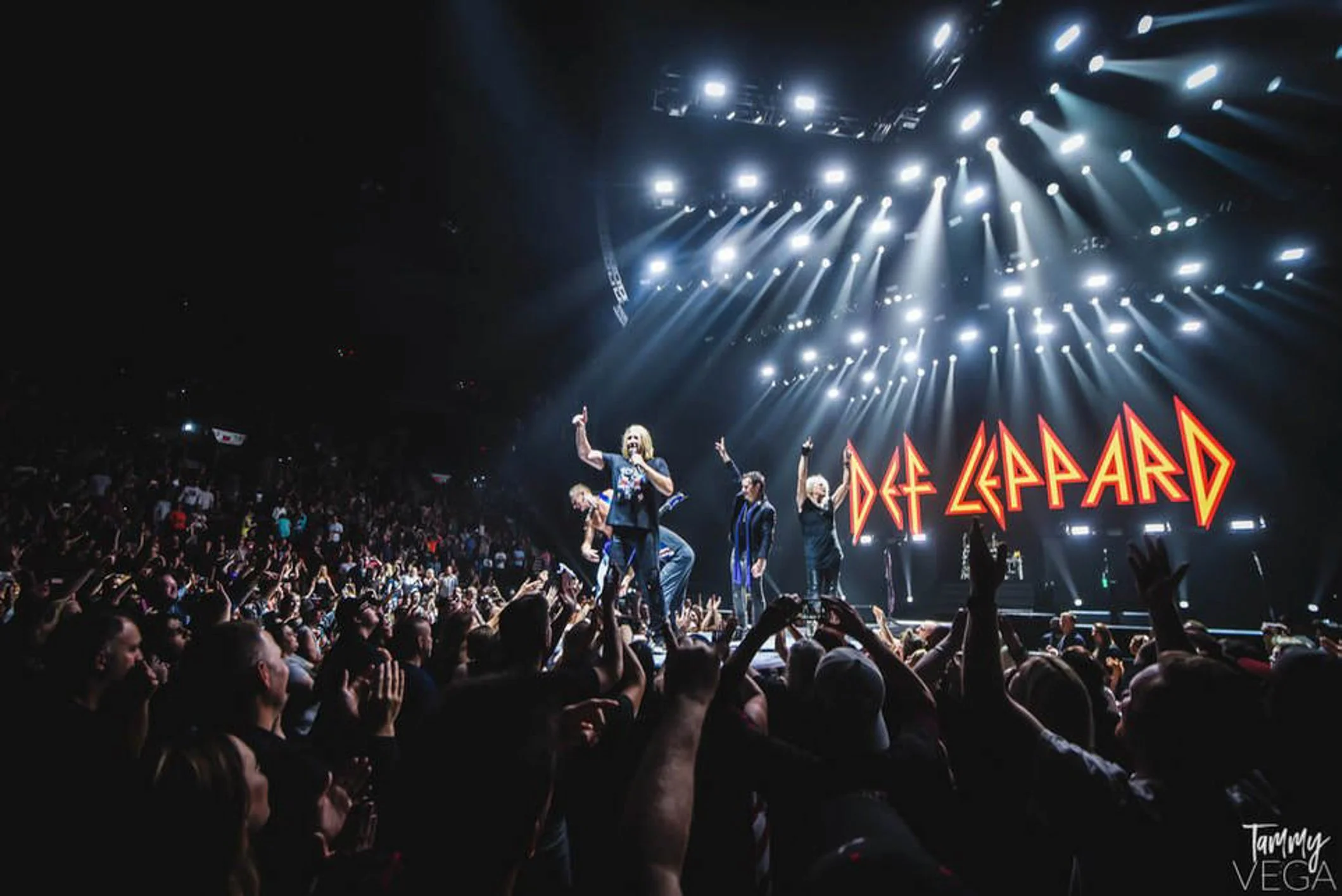 A band performing on stage during a concert, with bright stage lights and a large crowd in the audience.