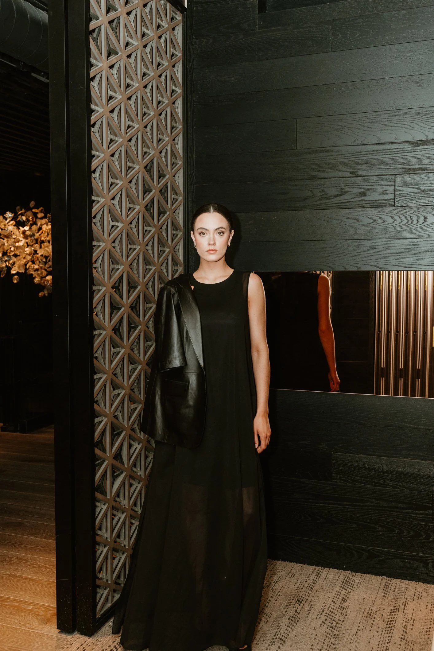 A woman with dark hair in a bun, wearing a black dress and black leather jacket, standing in a modern interior with black wood panels and a patterned wood partition.