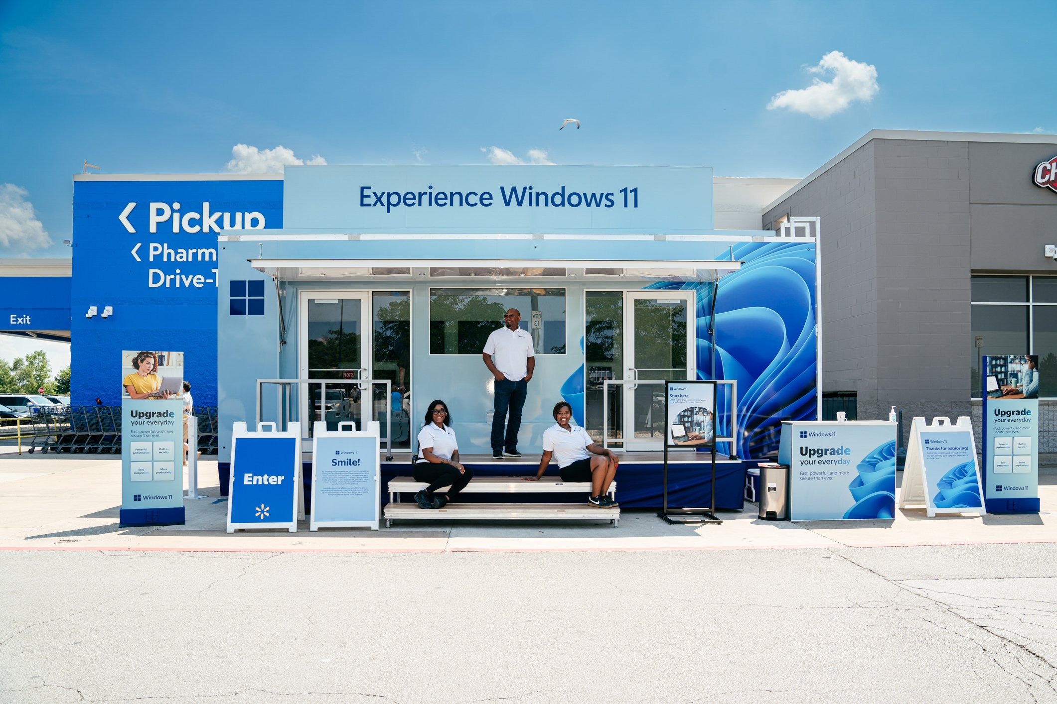 An outdoor promotional booth for Windows 11 with staff and visitors, featuring banners and signs, in a parking lot under a partly cloudy sky.