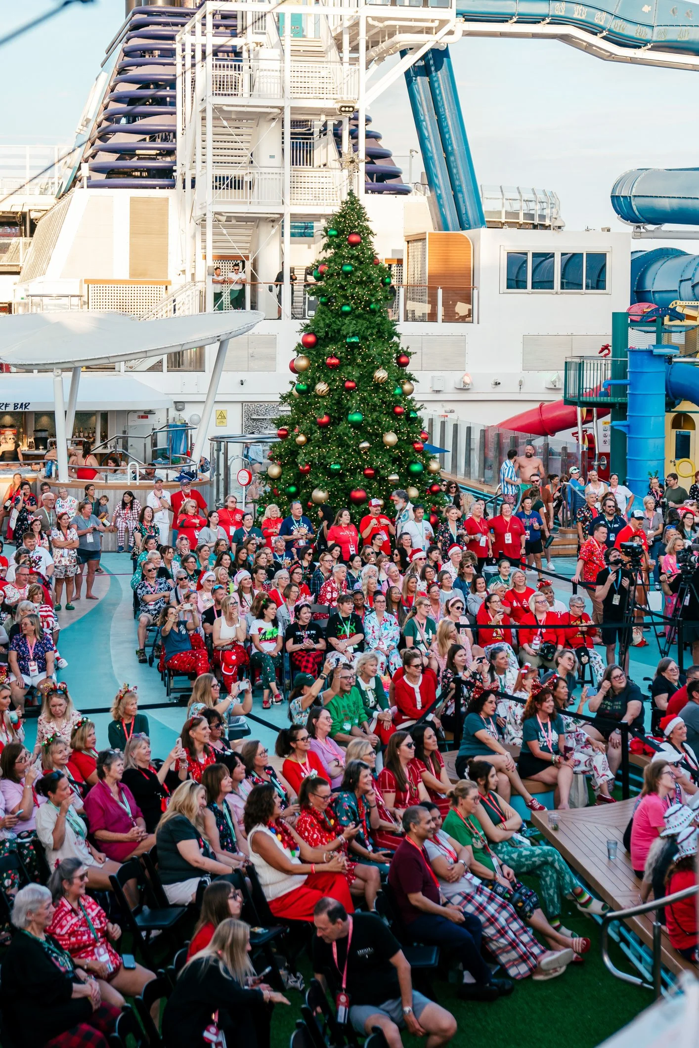 A large group of people gathered on a cruise ship deck for a Christmas celebration. There is a decorated Christmas tree in the center and water slides in the background. Many people are wearing holiday-themed clothing and festive accessories.
