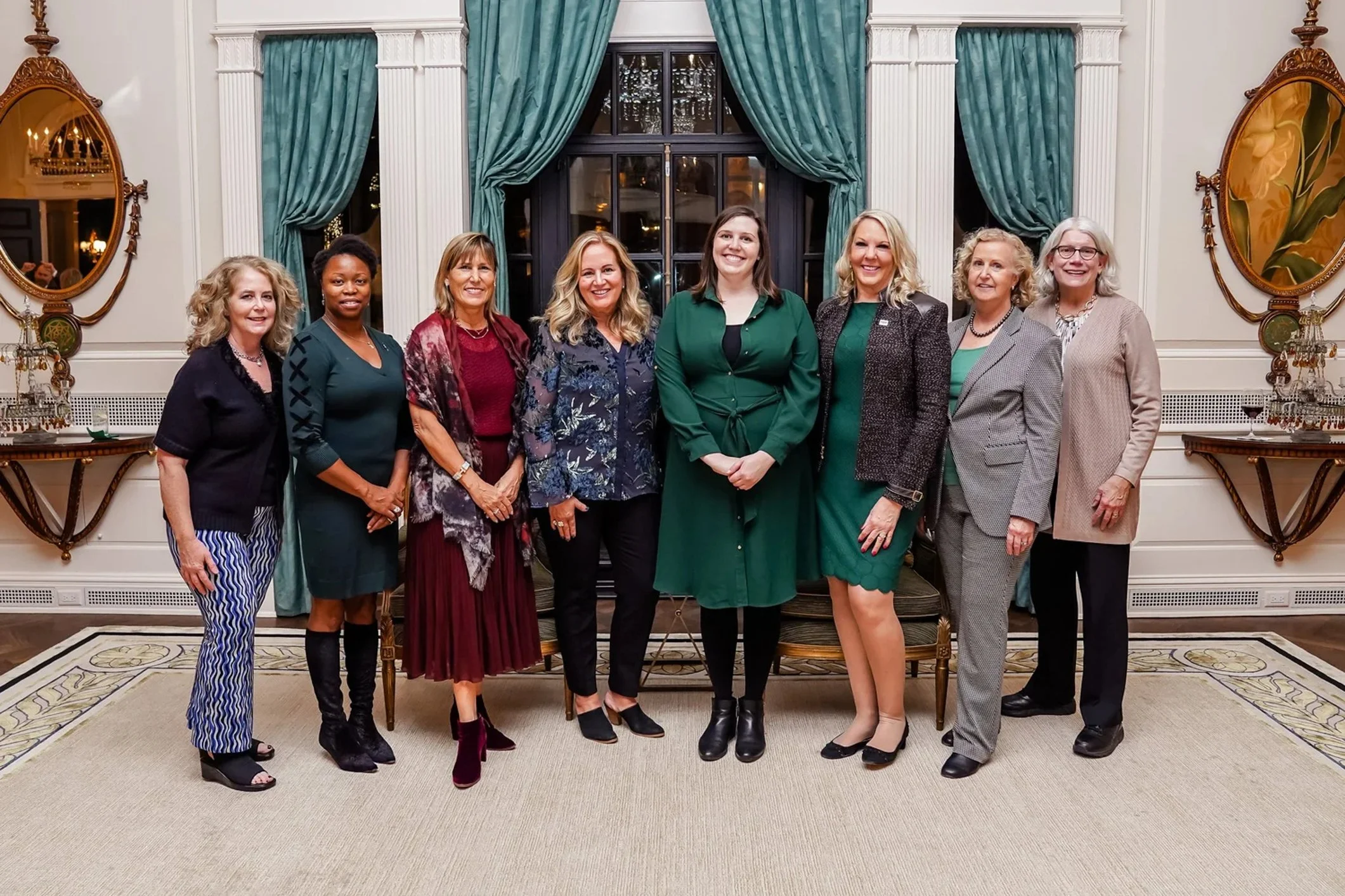 A group of nine women standing in a line inside a decorated room with large windows, teal curtains, and ornate mirrors.