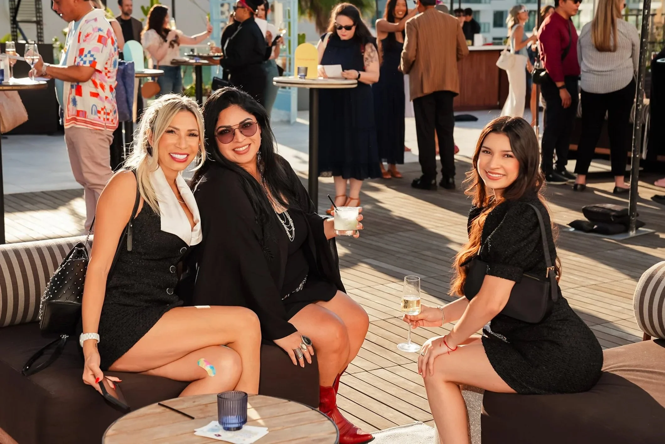 Three women sitting on a patio, smiling at the camera with drinks in hand at an outdoor social gathering in the late afternoon.