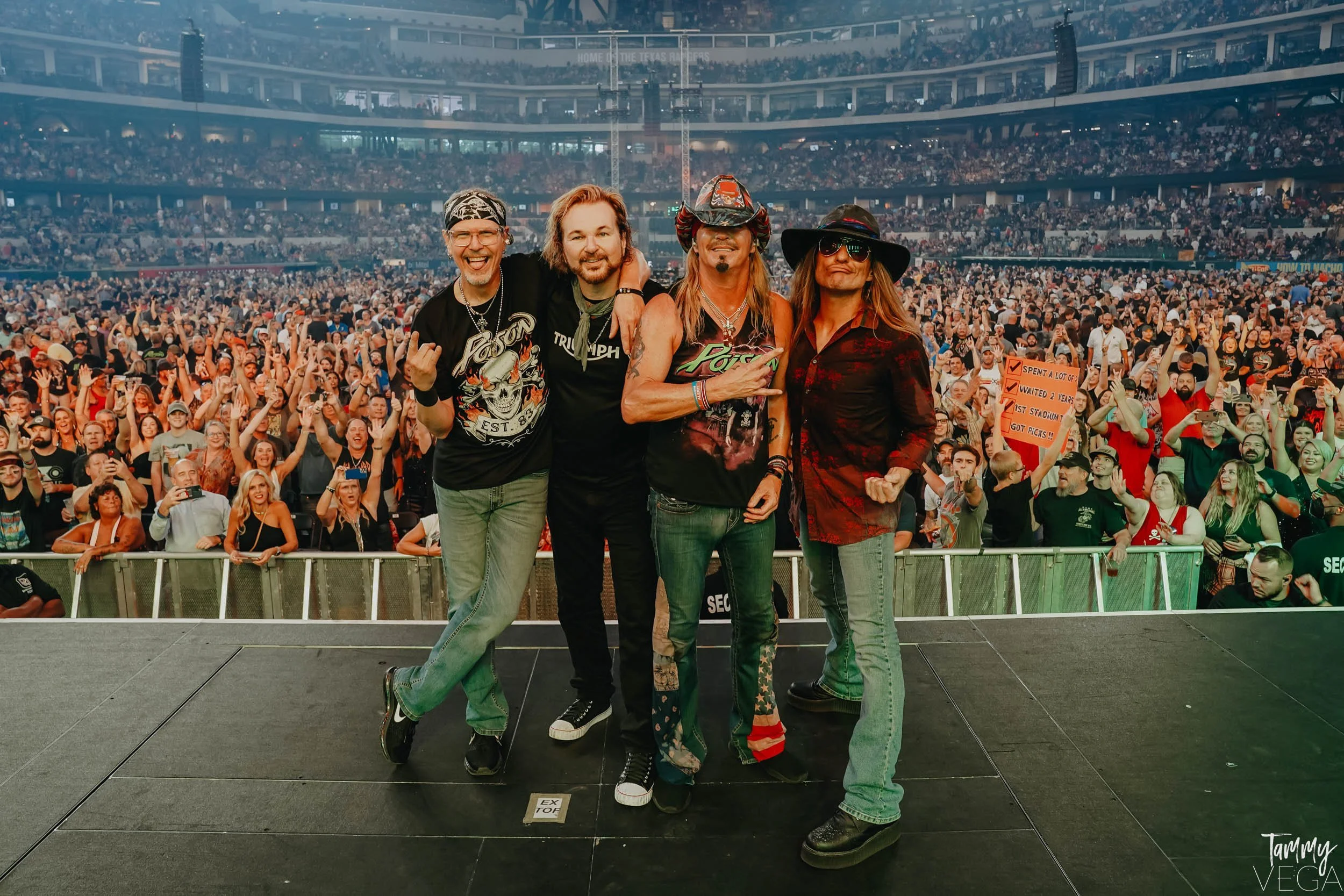 Four people standing on a stage at a crowded concert, smiling and posing for a photo in front of a large audience in a stadium.