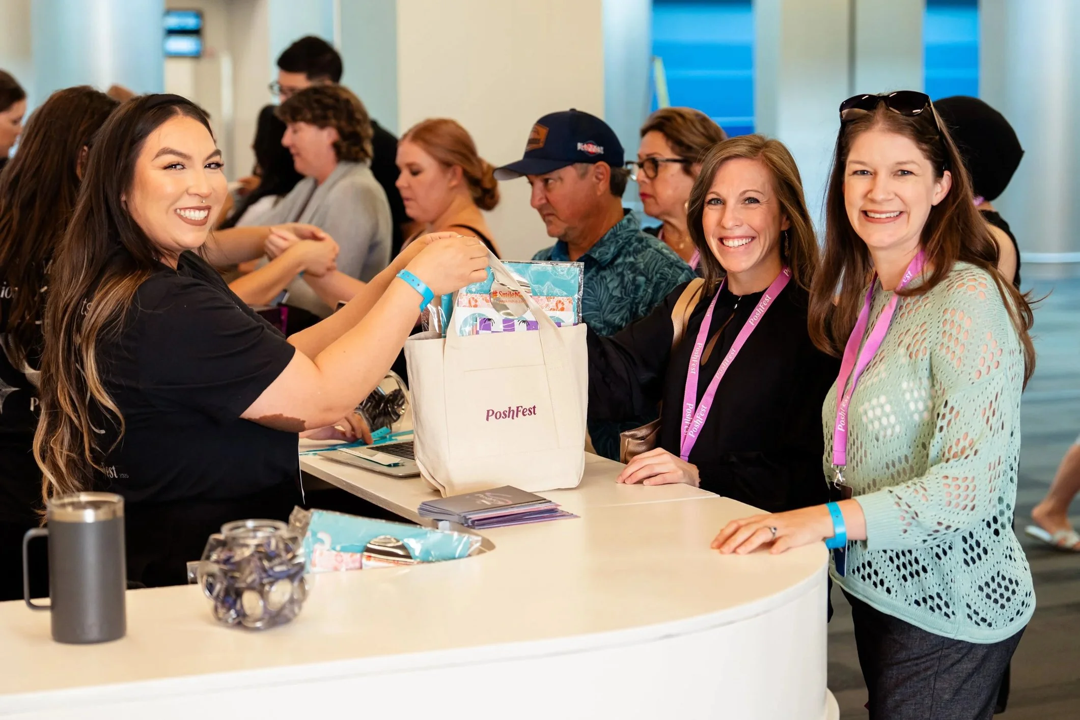 Smiling women at a registration table at Posh Fest, with other attendees in the background, some wearing lanyards and glasses.