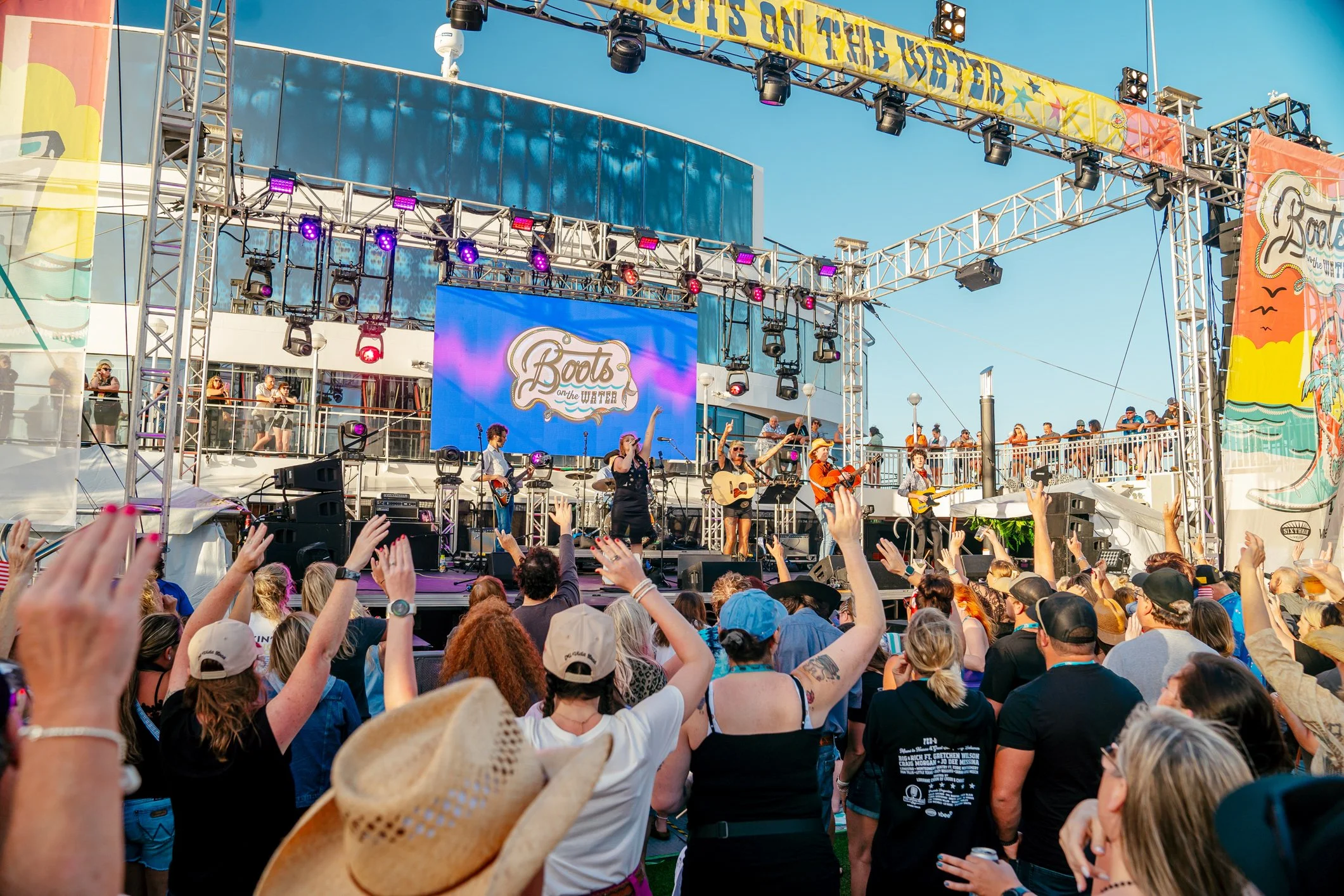 A live outdoor concert on a cruise ship with the sign 'Boots on the Water' on the stage. A band is performing with people dancing and raising their hands in front of the stage. The sky is clear and blue.