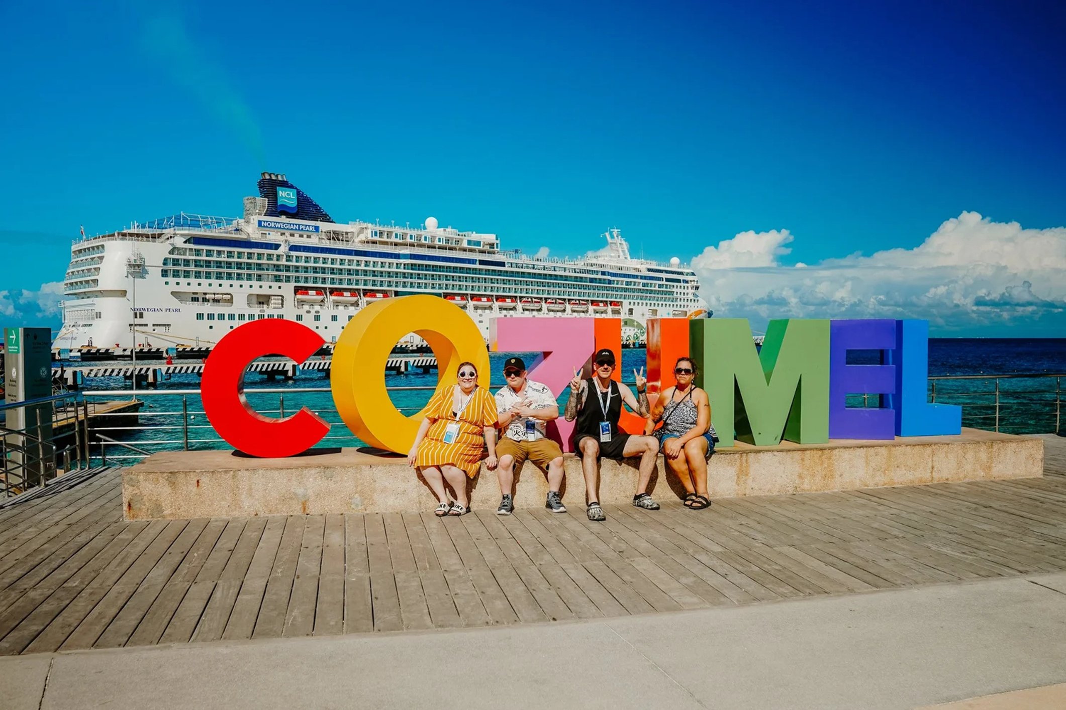 People posing in front of colorful letters spelling 'COSTA' at a sunny waterfront with a large cruise ship in the background.