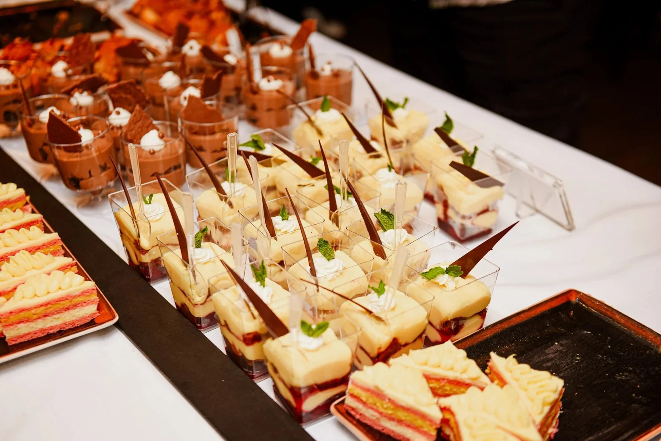 Assorted small desserts and cakes arranged on a display table, including chocolate mousse cups with whipped cream and chocolate decorations, and slices of layered cake with cream and fruit.