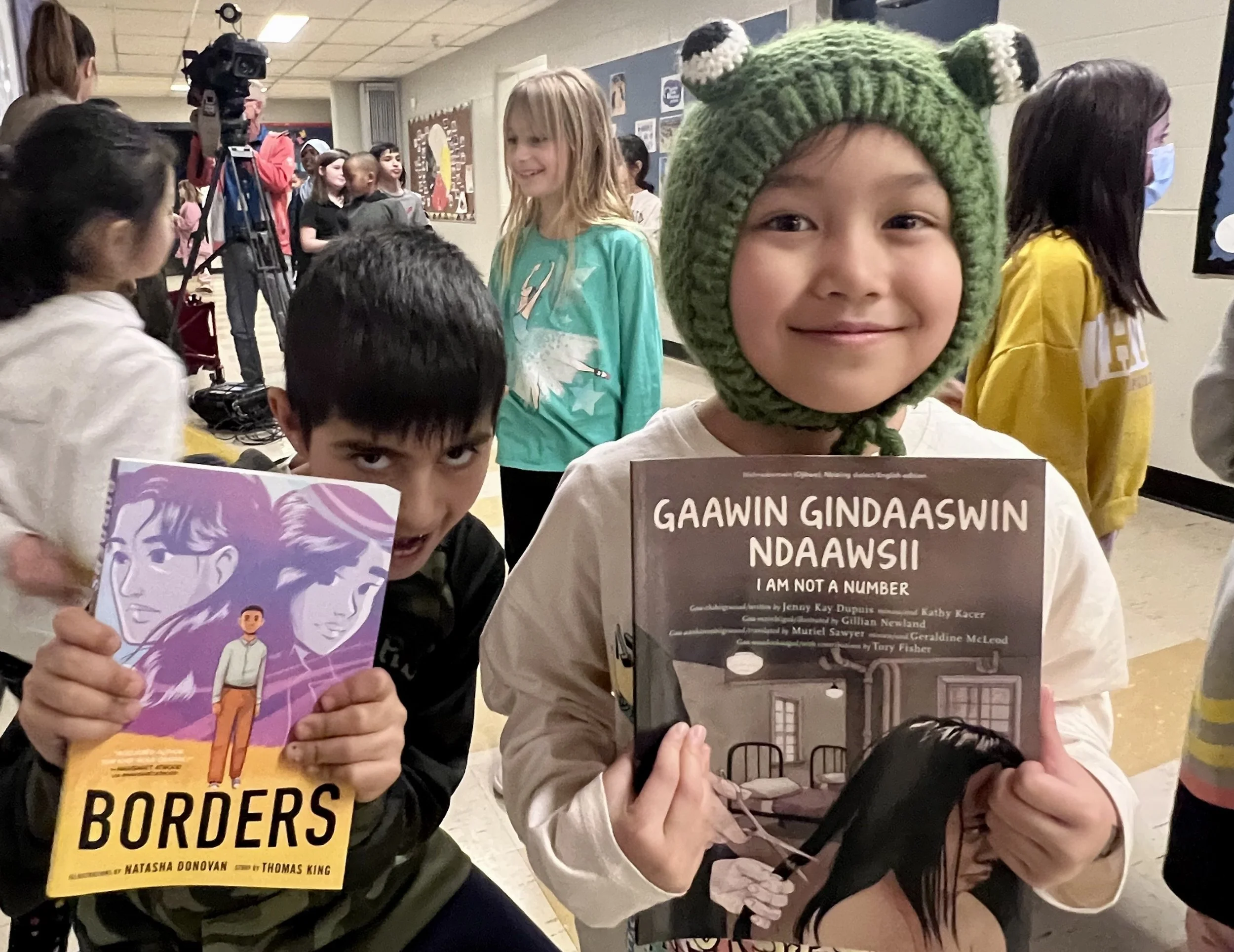 Children at a book event holding books, with some smiling and wearing casual clothing, in an indoor space with bulletin boards on the wall.
