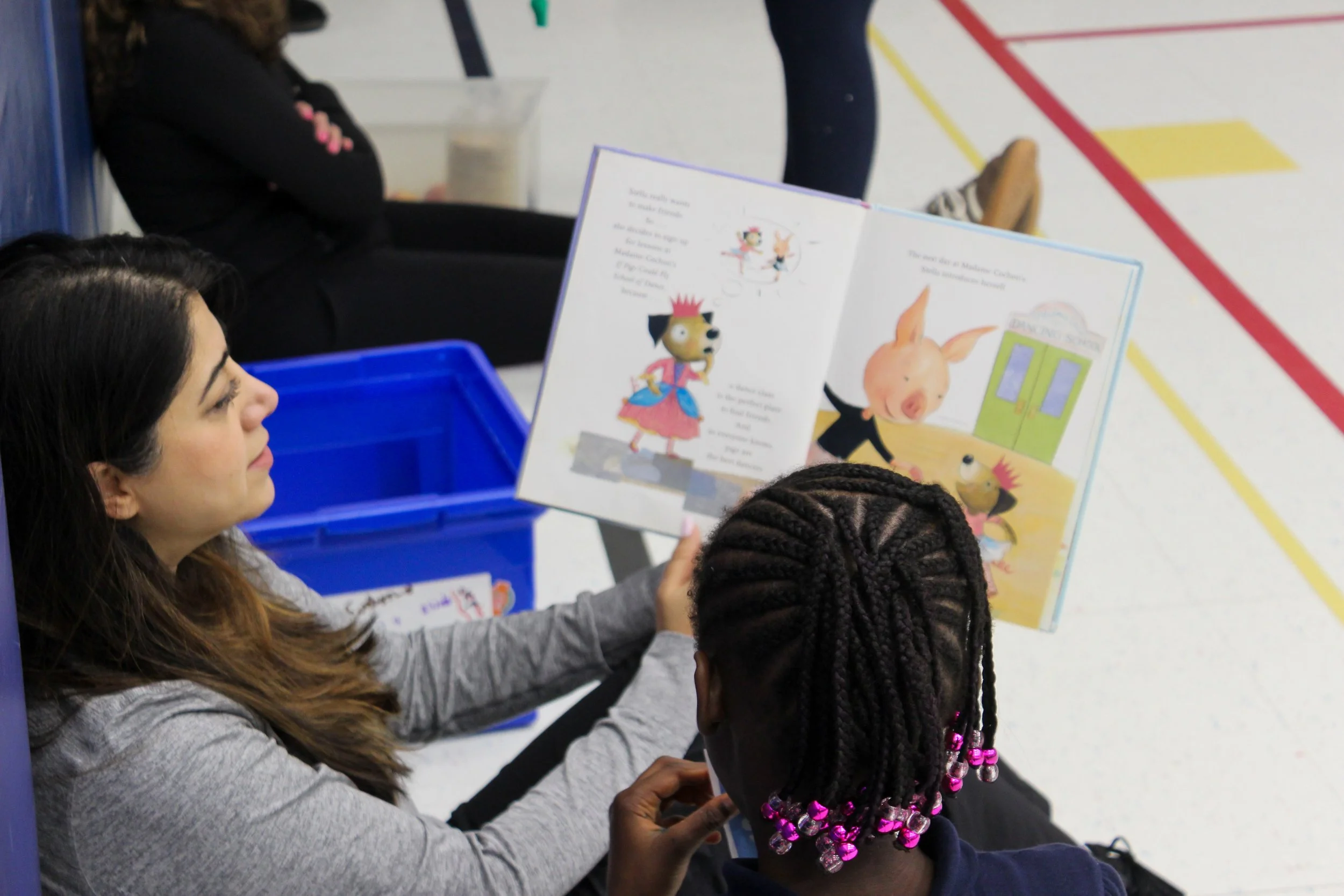 Children and an adult sitting at a table, reading a picture book with cartoon characters, in a gymnasium or school gym, with sports court markings on the floor.