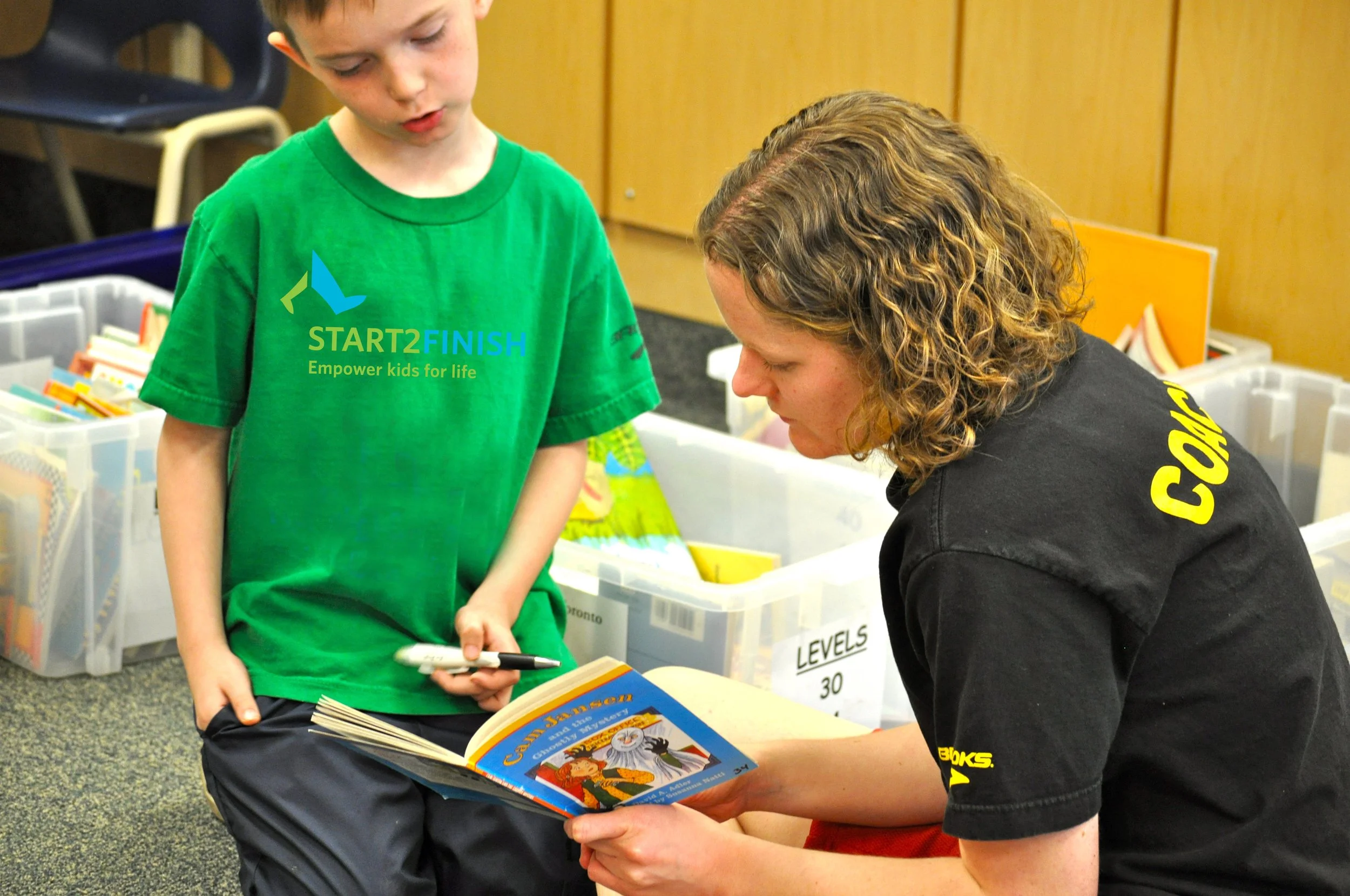 A young boy in a green t-shirt and a woman with curly hair in a black shirt are looking at a colorful children's book together in an indoor setting with storage bins and shelves around them.