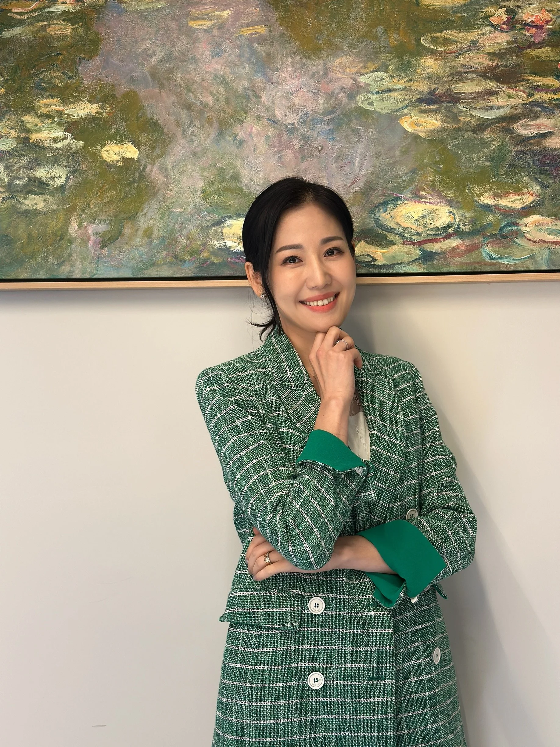 A smiling woman with black hair, wearing a green and white checkered blazer and skirt, standing in front of a large impressionist style painting of water lilies.