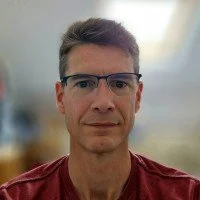 A man wearing glasses and a maroon shirt looking at the camera inside a well-lit room.