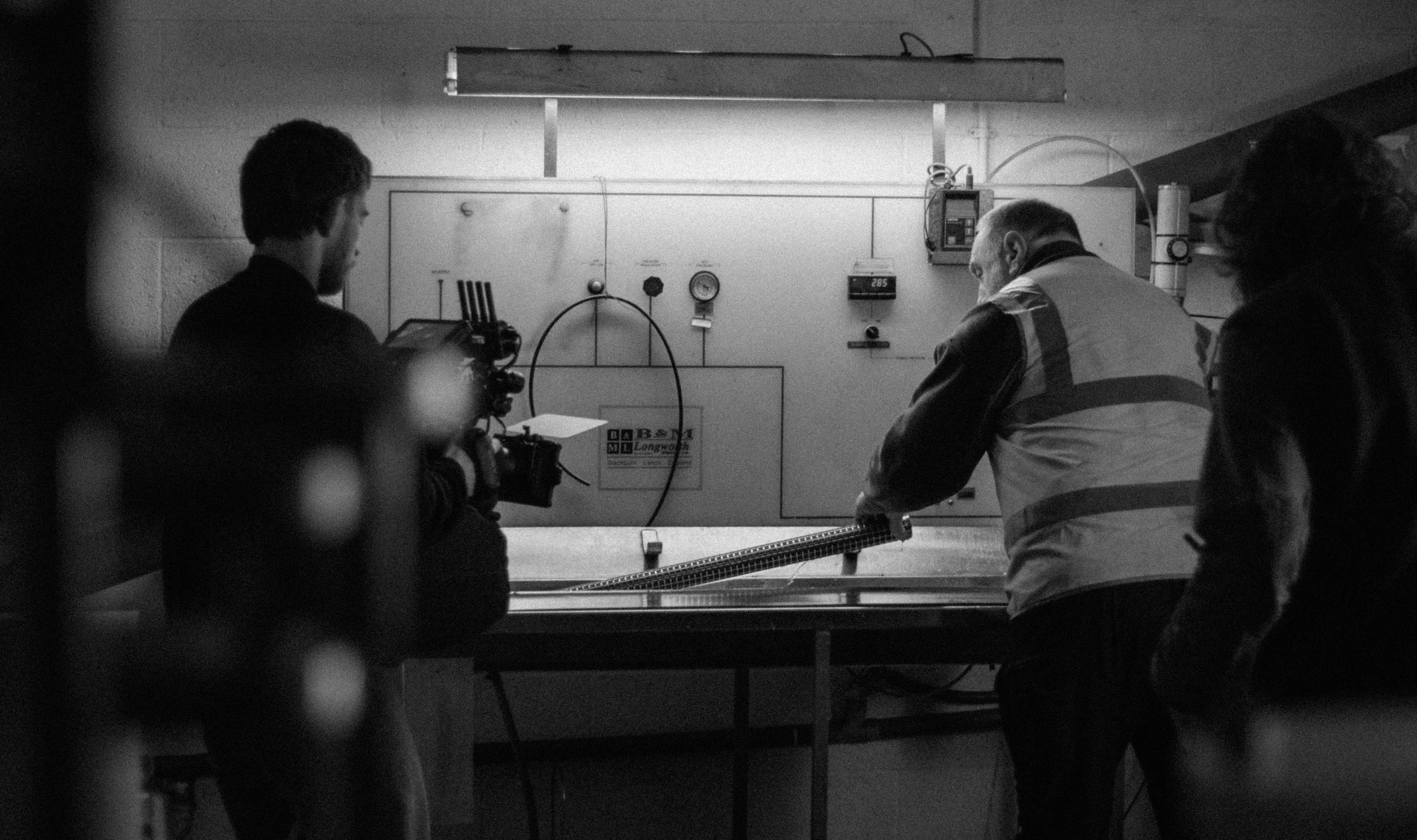 Black and white photo of three men in a room with technical equipment. One man is holding a camera, and another is looking at a wall-mounted control panel with gauges and devices. The third man is on the right side, partially visible, with long wavy hair.