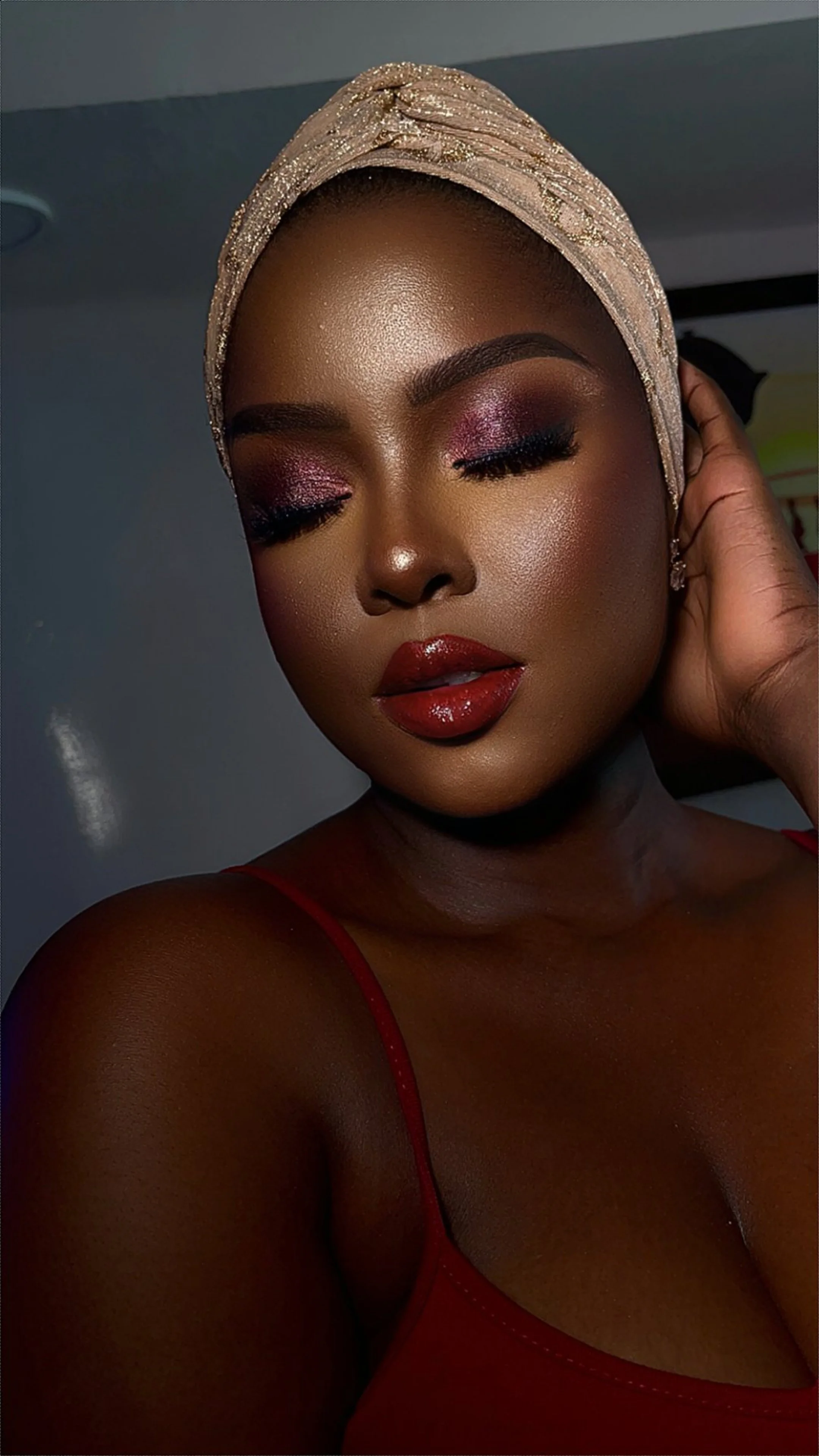 Close-up of a woman with dark skin, wearing a beige headwrap with gold details, red top strap, makeup with pink eyeshadow, long eyelashes, and dark red glossy lipstick, with one hand touching her head.