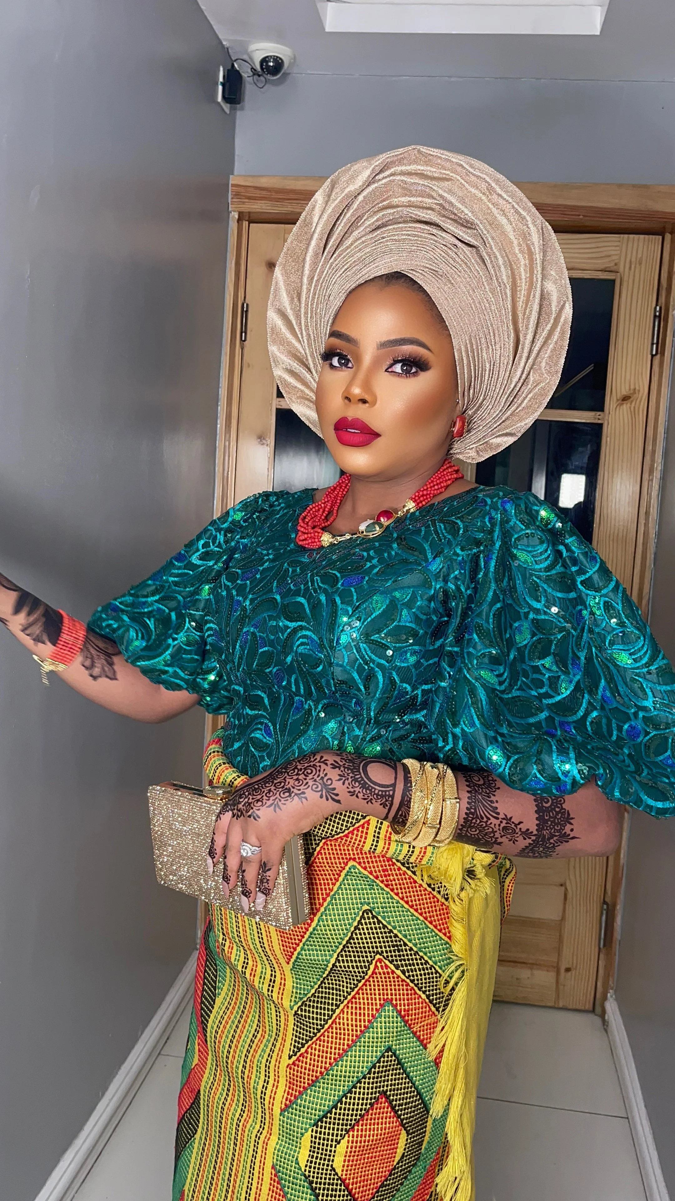 A woman dressed in traditional African attire with a large beige headwrap, earrings, necklaces, bangles, and intricate henna tattooed hands, standing indoors next to a wooden door.