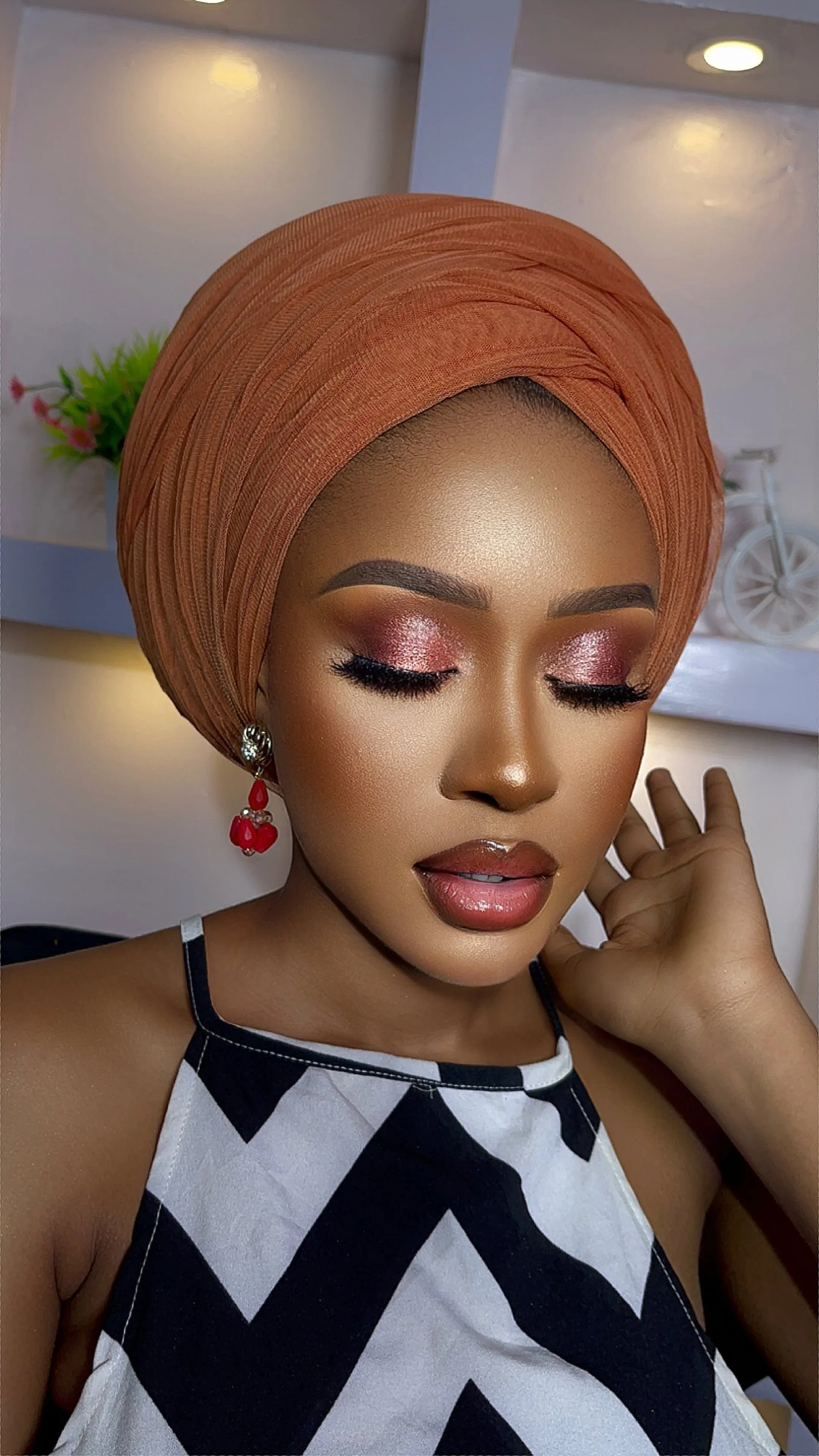 A woman with makeup and earrings wearing an orange headwrap and a black and white patterned top, with a blurred background featuring decor and a white decorative bicycle.