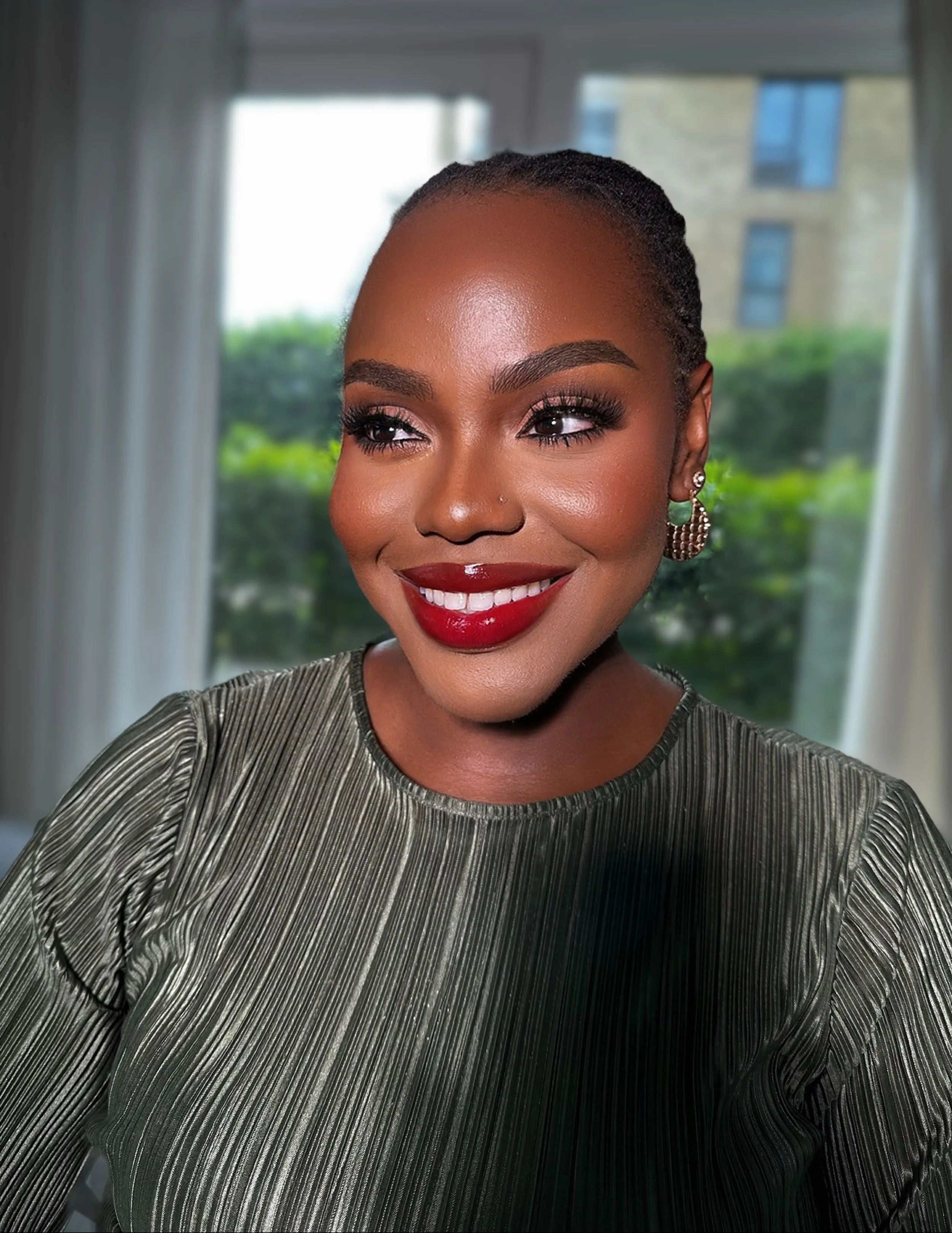 A smiling woman with short hair, red lipstick, and earrings, wearing a ribbed metallic top, indoors with a window showing greenery outside.