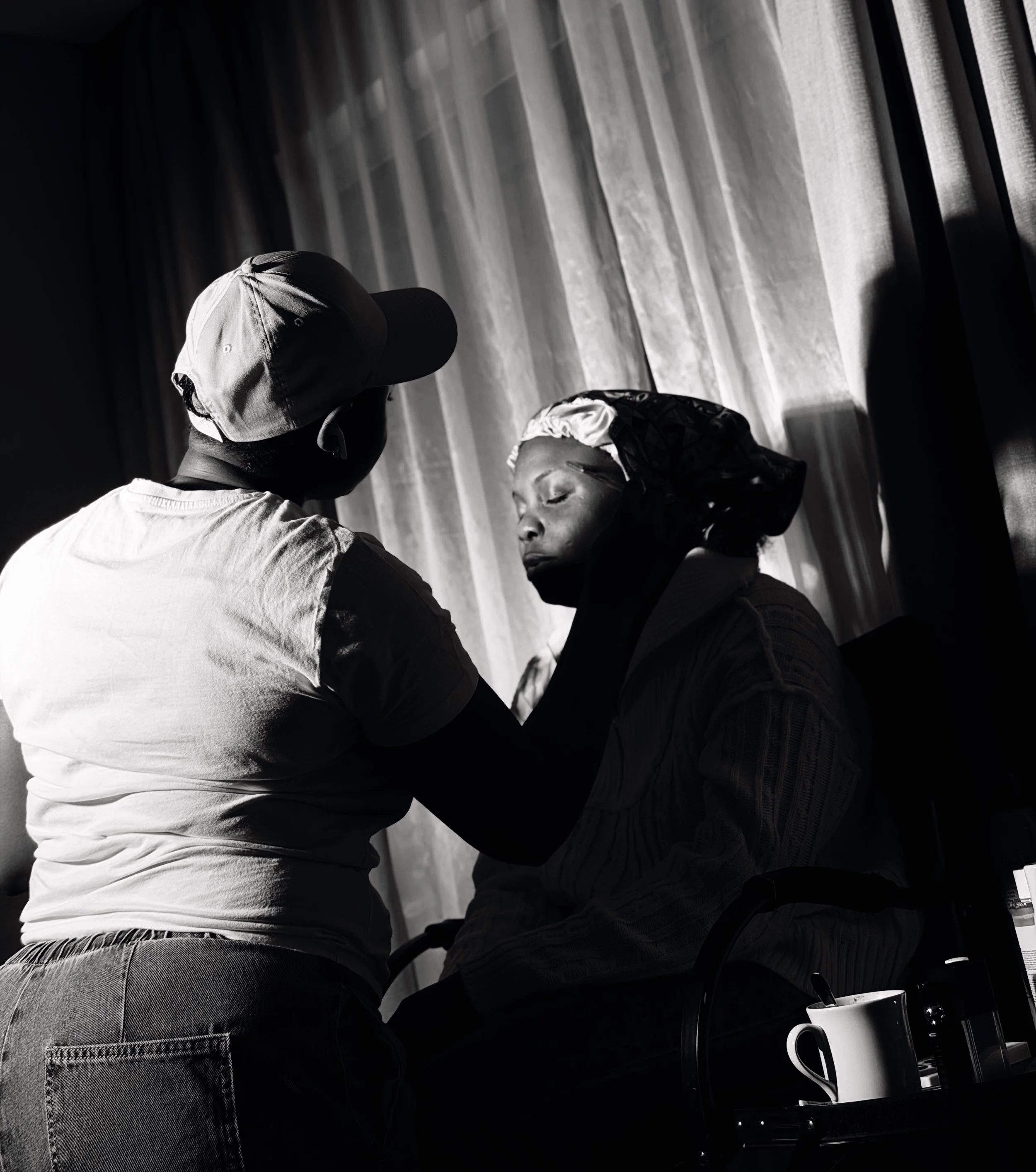 A black-and-white photo of two women sitting close together, facing each other, engaged in conversation. One is wearing a cap and a face mask, while the other wears a headscarf. The setting appears to be indoors, with vertical wooden or panel walls in the background.