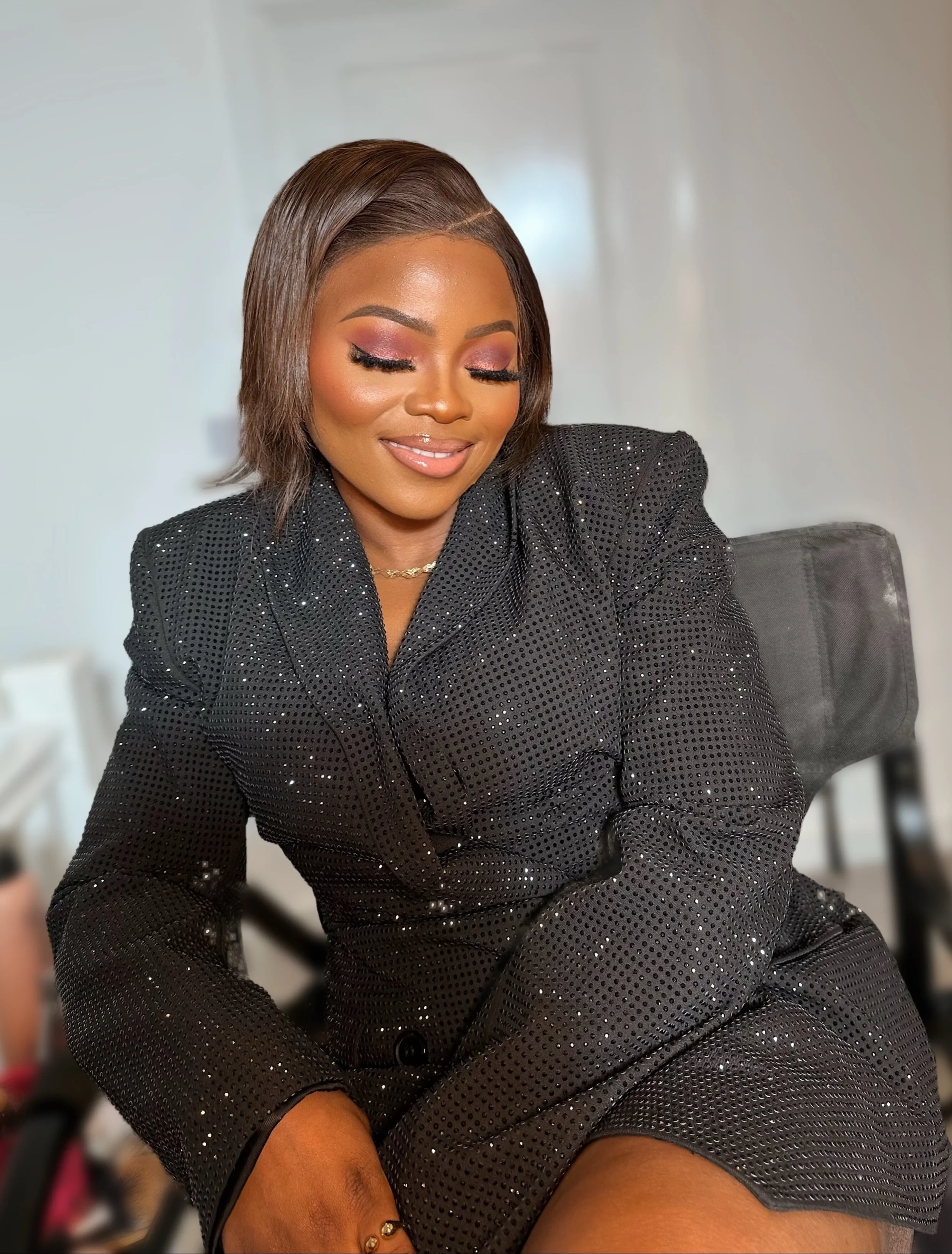A woman with shoulder-length brown hair wearing a black sparkly blazer and makeup, sitting with her eyes closed and smiling.
