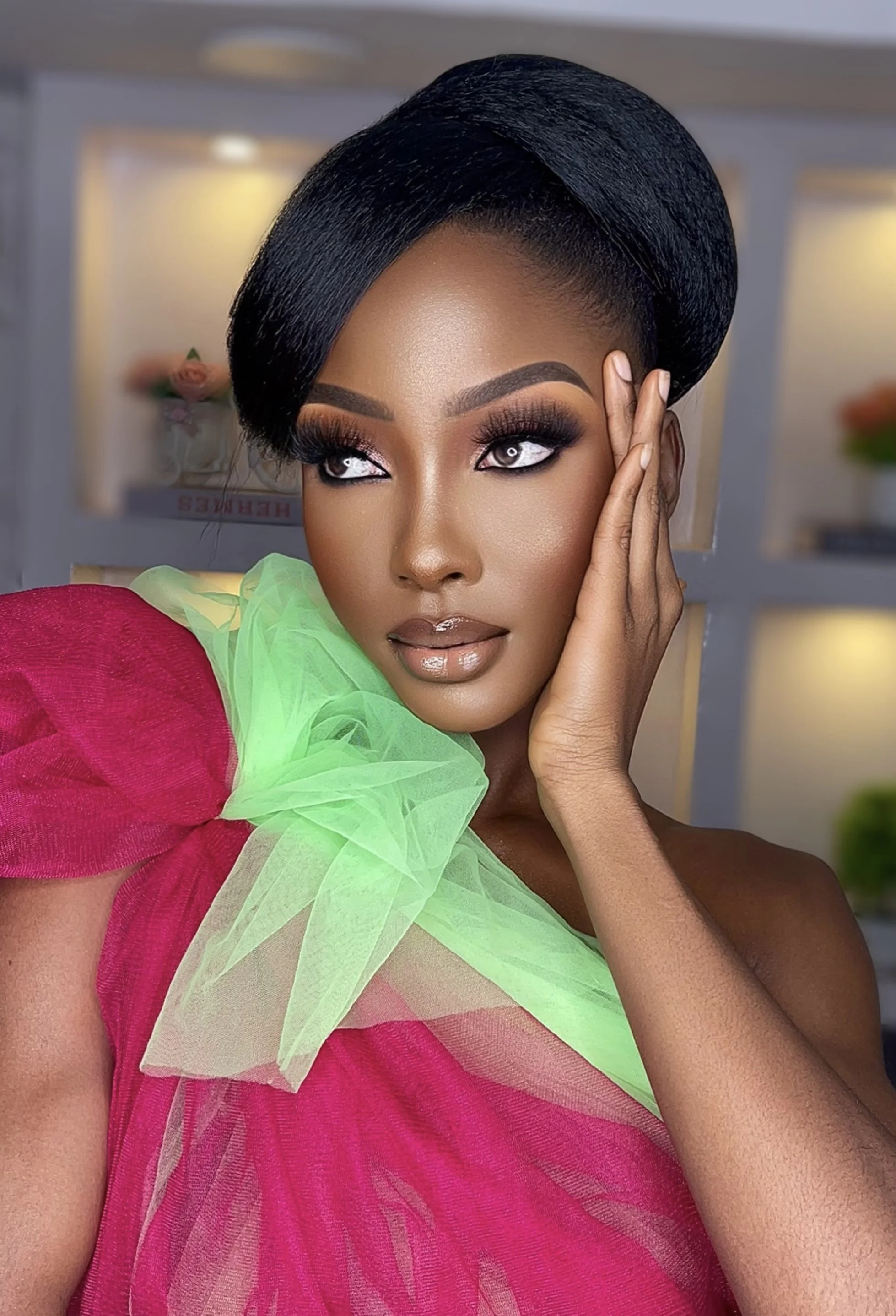 A woman with dark, styled hair, bold makeup, and glossy lips wearing a bright pink dress with green tulle accents poses indoors.