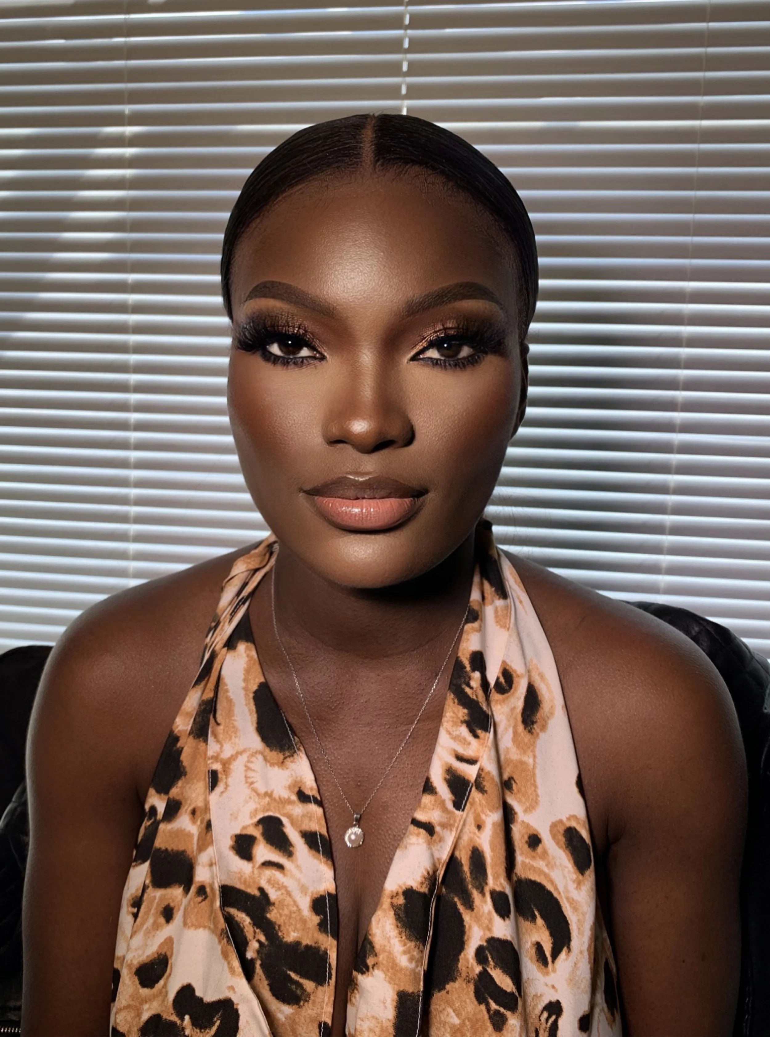 A woman with dark skin, sleek hair, and dramatic makeup, wearing a leopard print top and a silver necklace with a heart-shaped pendant, standing in front of closed white blinds.