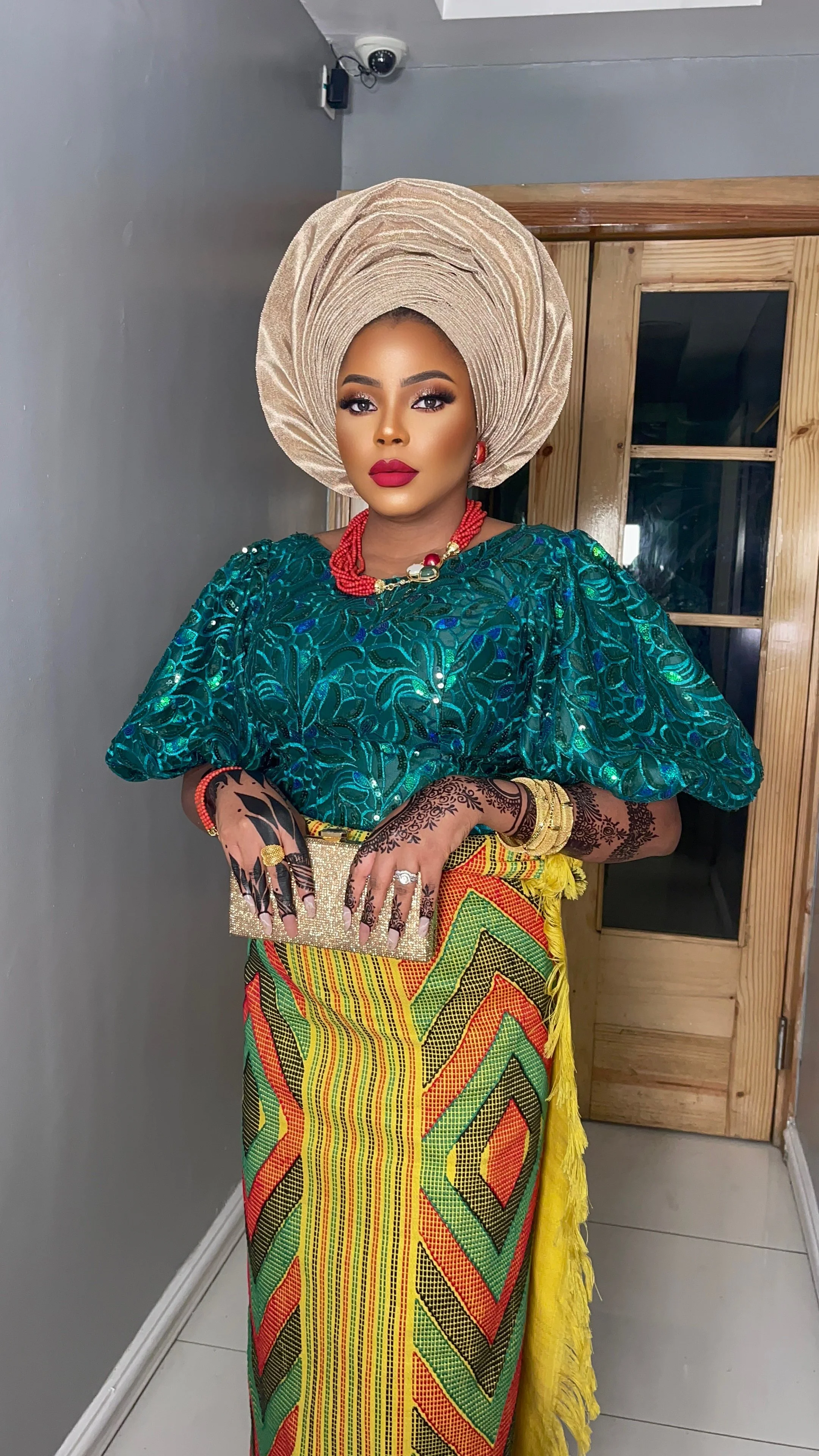 A woman dressed in traditional Nigerian attire, wearing a large beige gele headwrap, a vibrant green and blue lace top, and a colorful patterned wrapper skirt. She has henna on her hands, jewelry including a red beaded necklace, earrings, and gold bangles, and holds a glittery gold clutch. She stands indoors next to a wooden door.