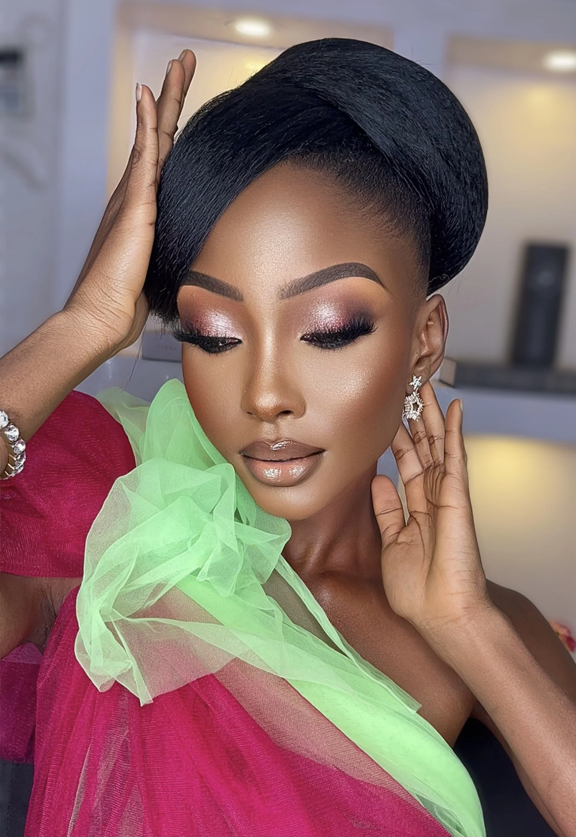 Close-up of a woman with makeup wearing a black hairstyle and earrings, with one hand on her face and the other touching her hair, dressed in vibrant pink and green fabrics.
