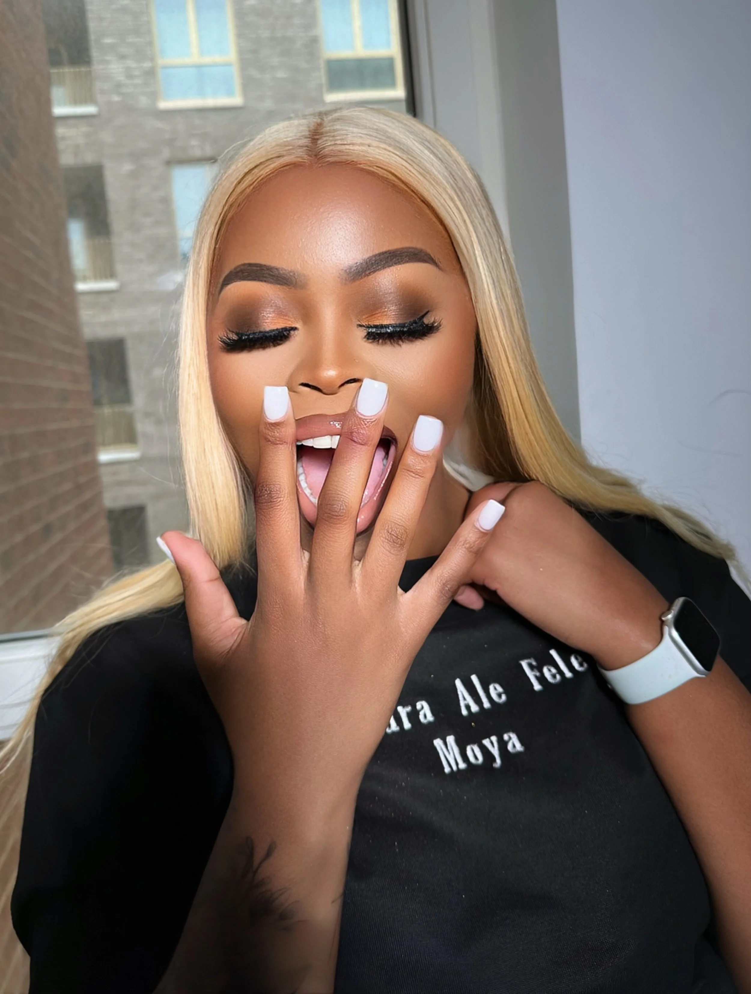 A woman with long blonde hair, closed eyes with makeup, and white nails, is making a peace sign near her mouth with her tongue slightly out, inside a room with a window showing a building outside. She is wearing a black top with white text and a whit