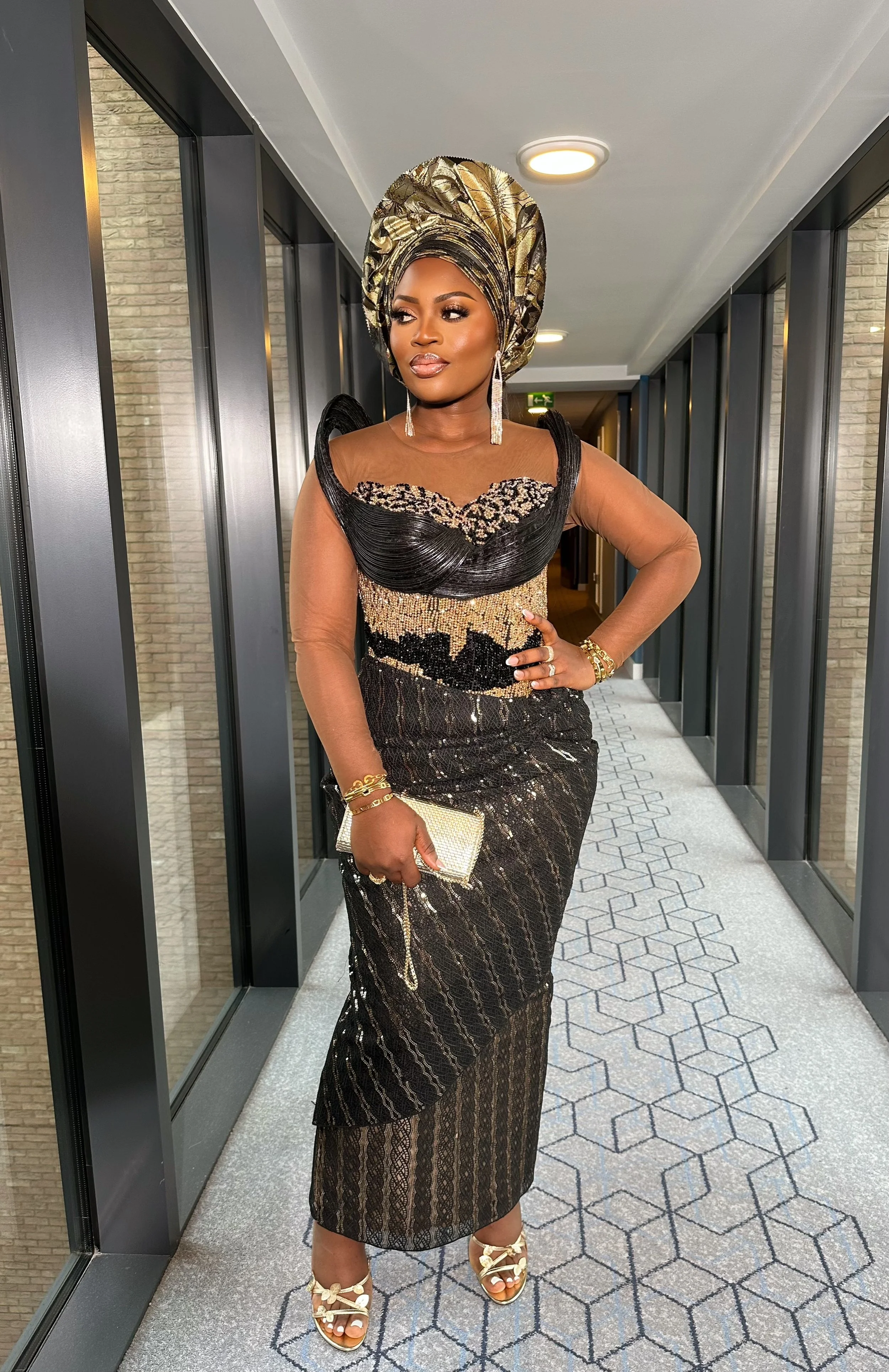 A woman in an elegant black and gold dress with intricate patterns, standing in a well-lit corridor with mirrors and brick walls, wearing a gold and black headwrap, jewelry, and heels, holding a gold clutch.