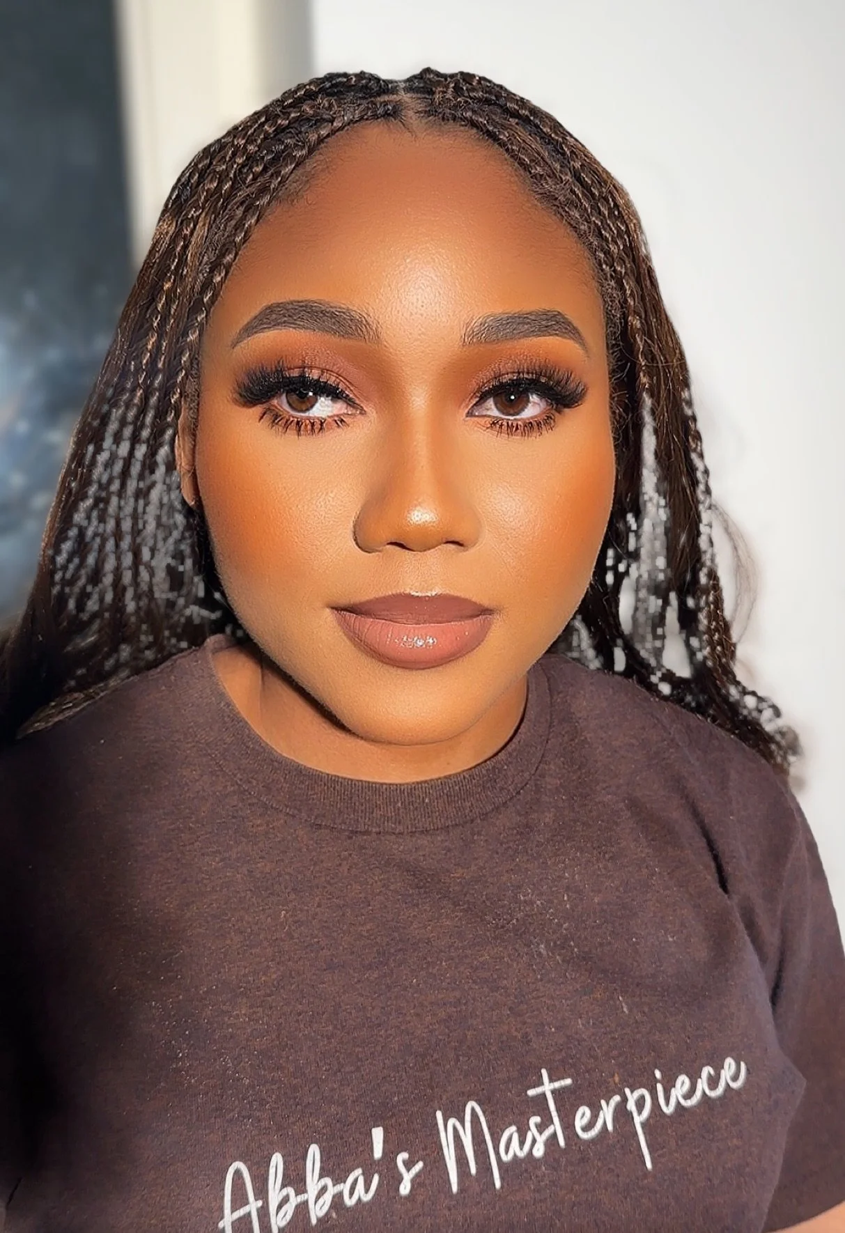 A young woman with long braids, makeup, and a brown shirt with white text. Her makeup includes brown eyeshadow, false eyelashes, and glossy lipstick.