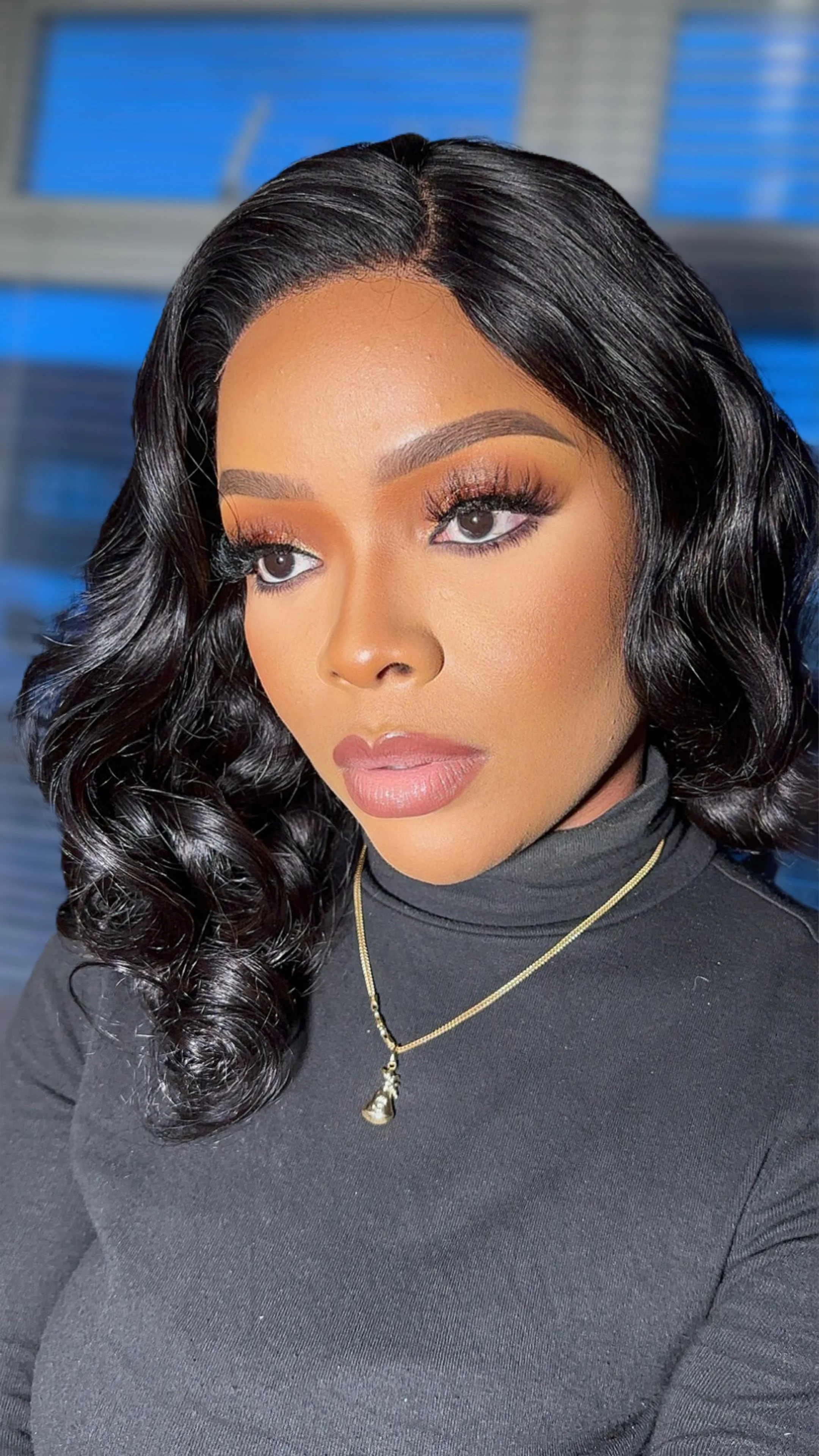 Close-up of a woman with curly black hair, wearing makeup with defined eyebrows, long eyelashes, and nude lipstick, dressed in a black top with layered necklaces, in an indoor setting with blue and gray background.