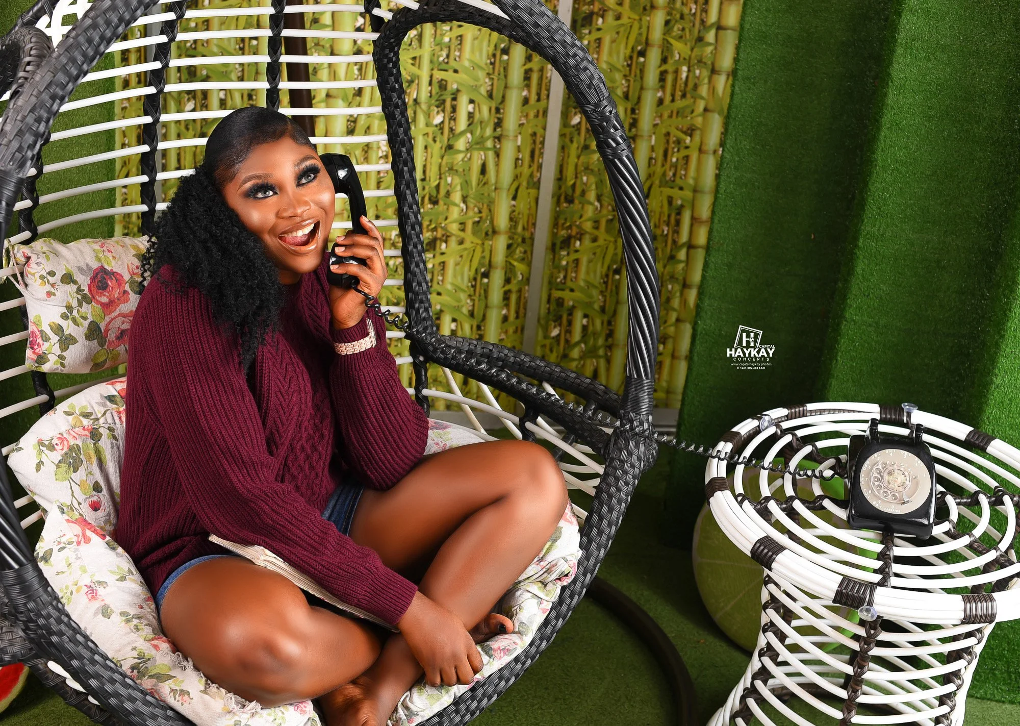 A woman with long braids, wearing a maroon sweater and shorts, sits on a floral cushion in a hanging wicker chair, talking on a black rotary phone, smiling happily. The scene is set outdoors with a backdrop of bamboo and a green artificial grass wall, next to a white wicker table and a vintage style alarm clock.