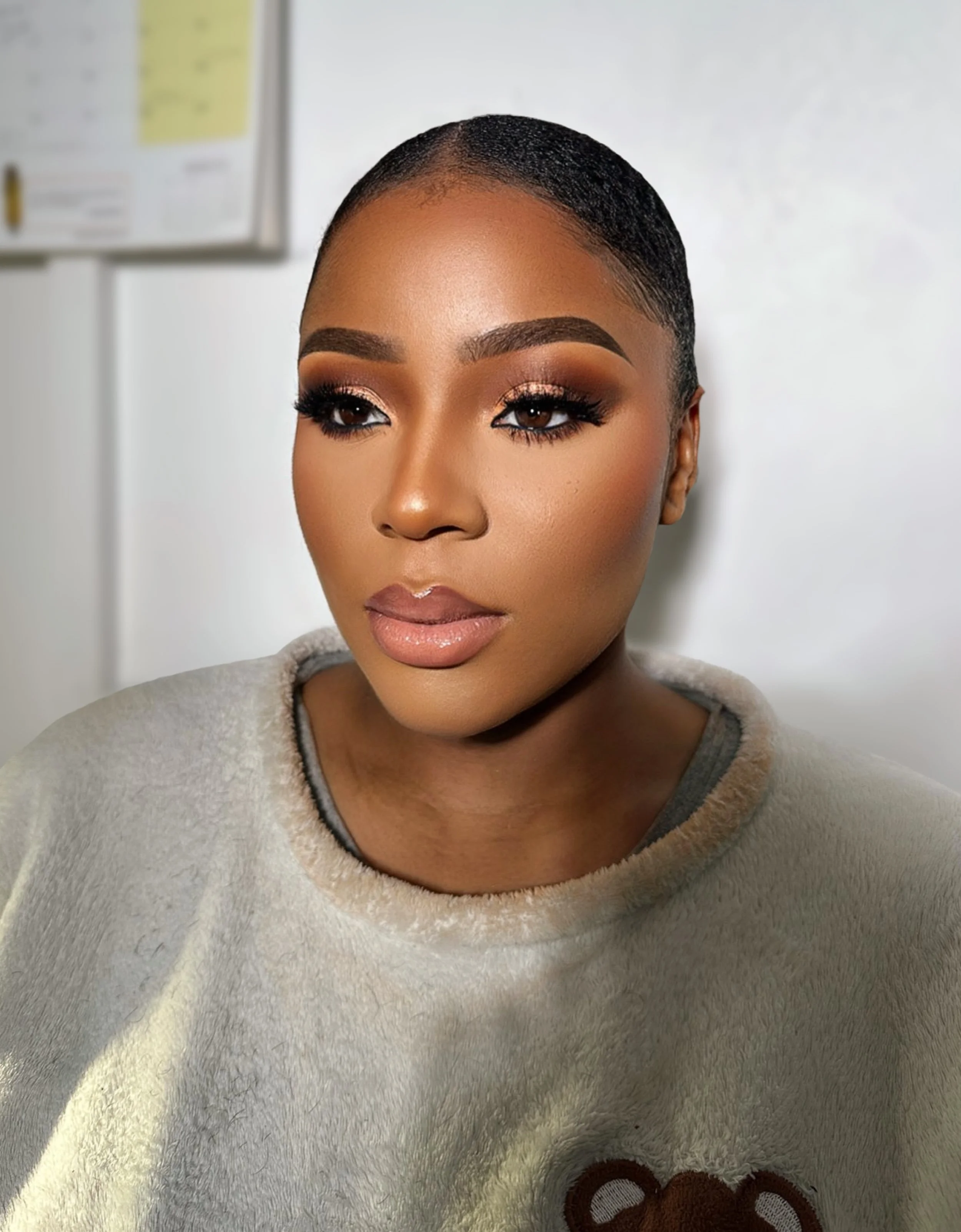 Close-up of a woman with short, sleek hair, well-defined eyebrows, and makeup, wearing a beige fleece sweater, in an indoor setting.