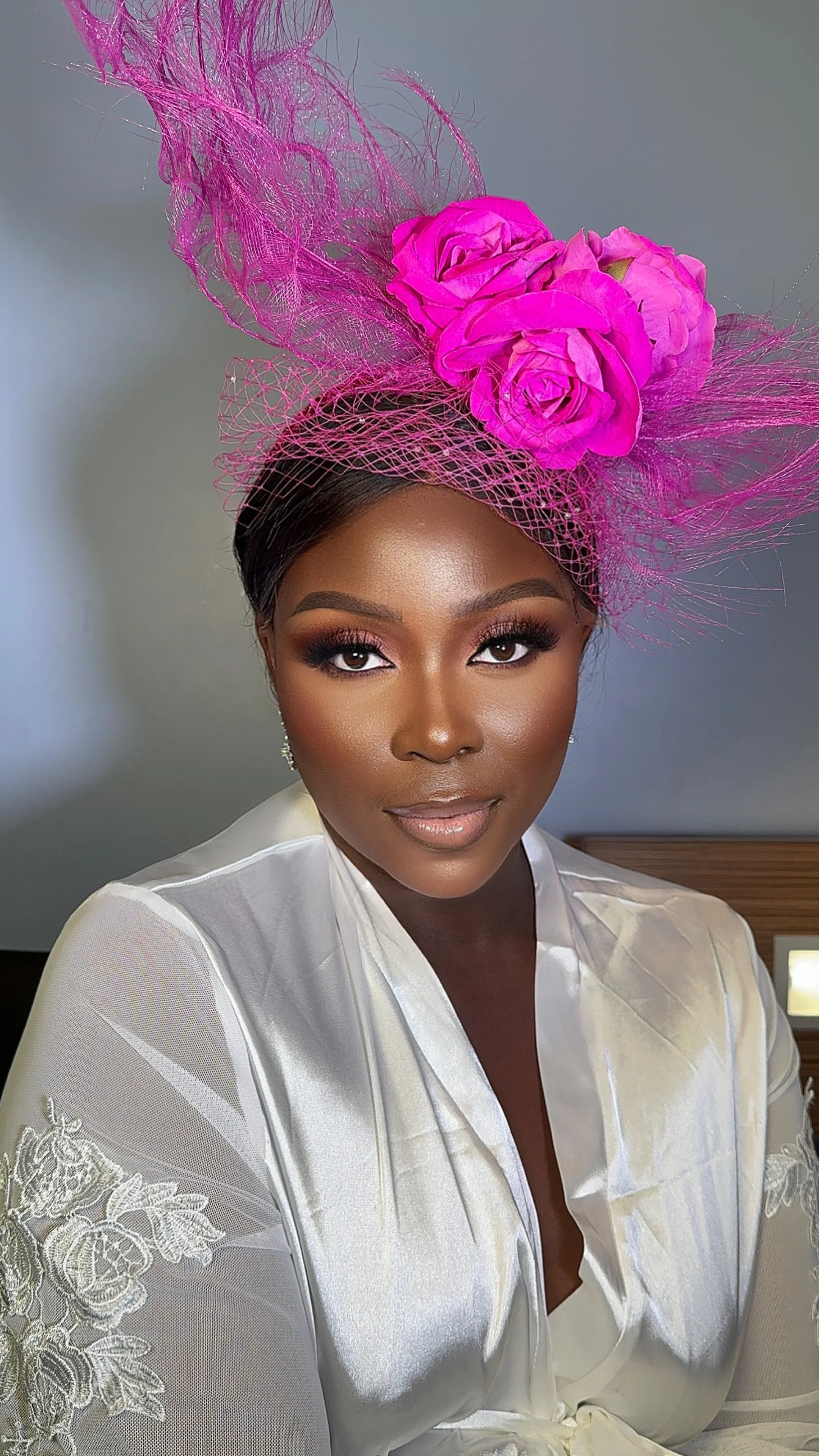 A woman with dark hair and makeup, wearing a white satin robe with lace embroidery, and a large pink floral headpiece with netting.