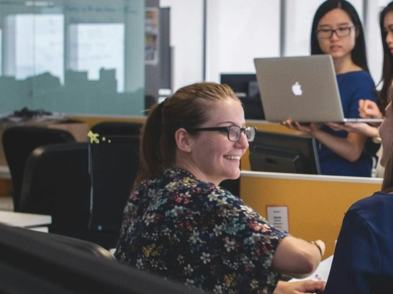 A woman with glasses and a floral shirt smiling and sitting in an office, with two women in the background working on laptops.