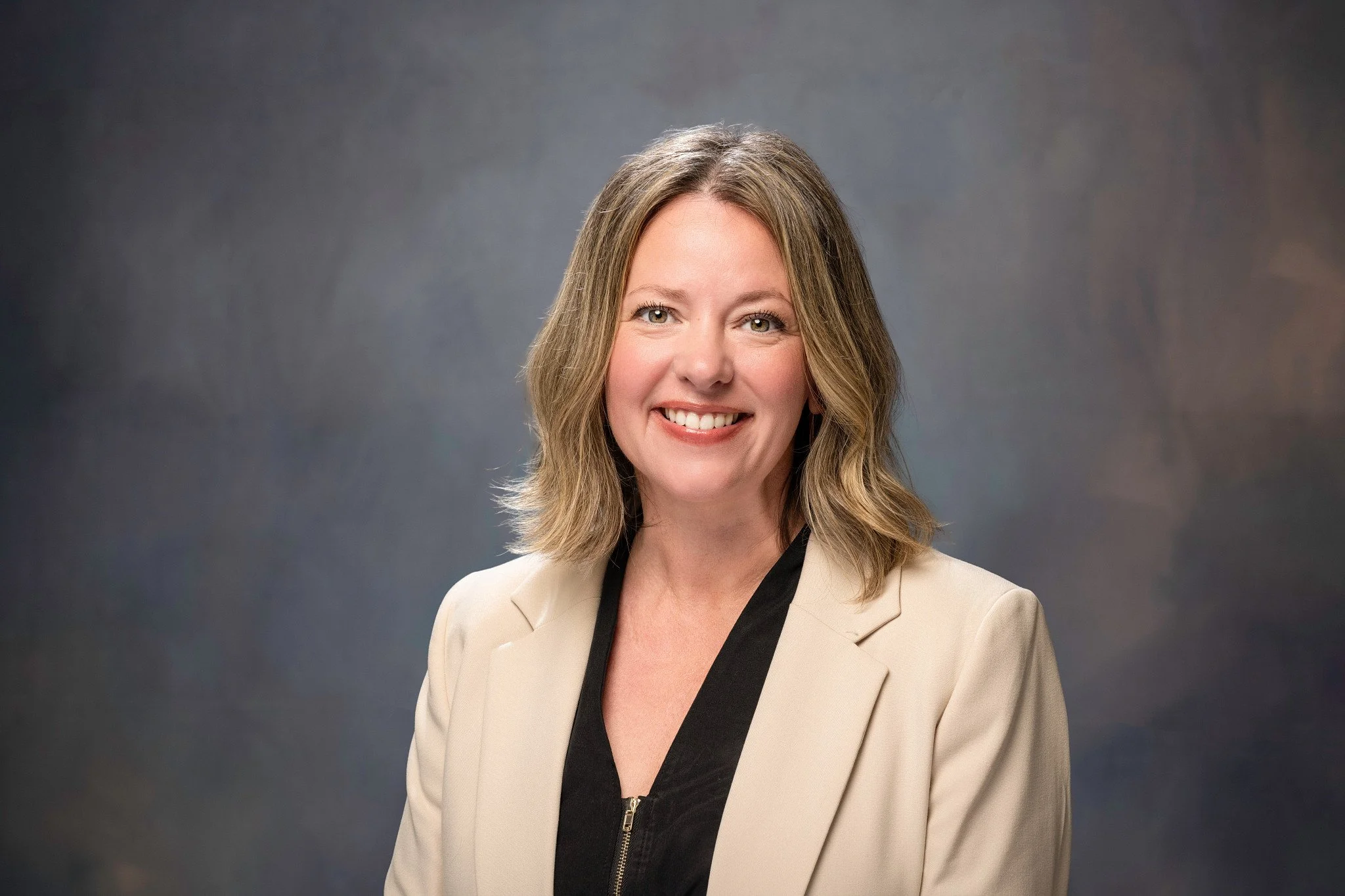 A woman with shoulder-length blonde hair and a friendly smile, wearing a beige blazer over a black top, against a dark gray background. Financial Advisor April Dwiggins.