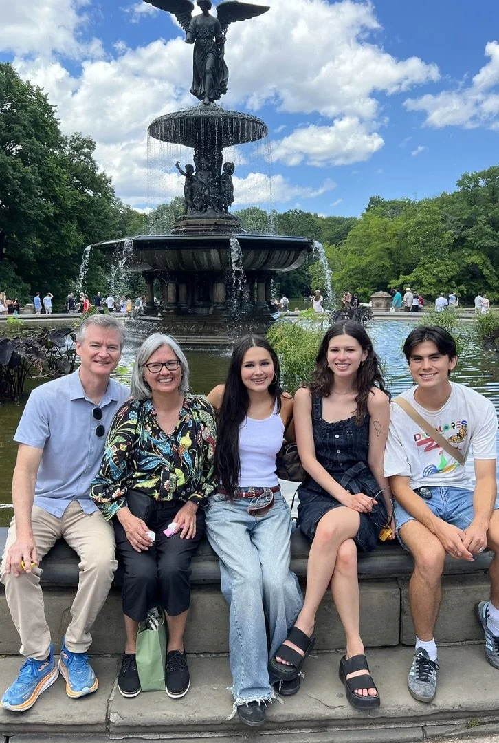 A group of five people sitting on a bench in front of Bethesda Fountain in Central Park, New York City, with the fountain and a crowd of people in the background during daytime.