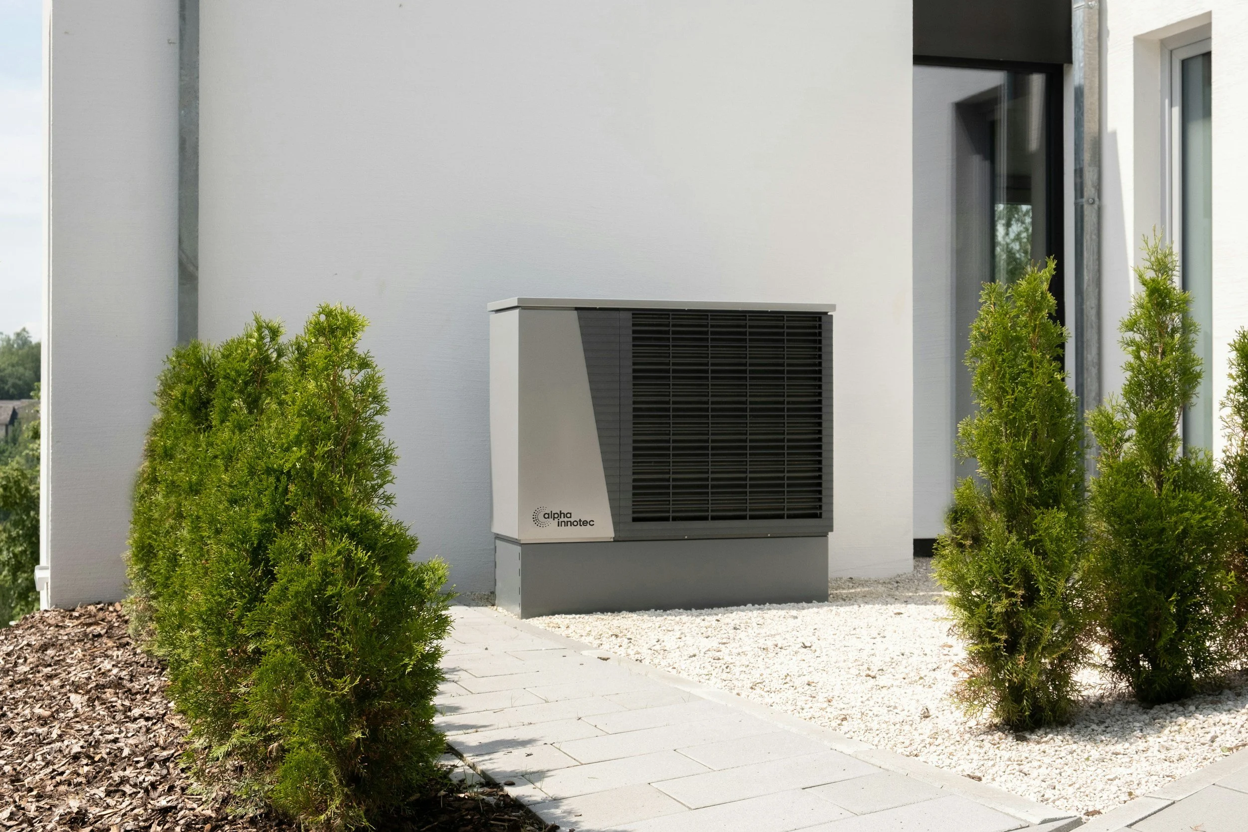 Exterior of a modern white house with a black-framed window and garden bushes, featuring a gray air conditioning unit branded Alpha Innotec nearby.