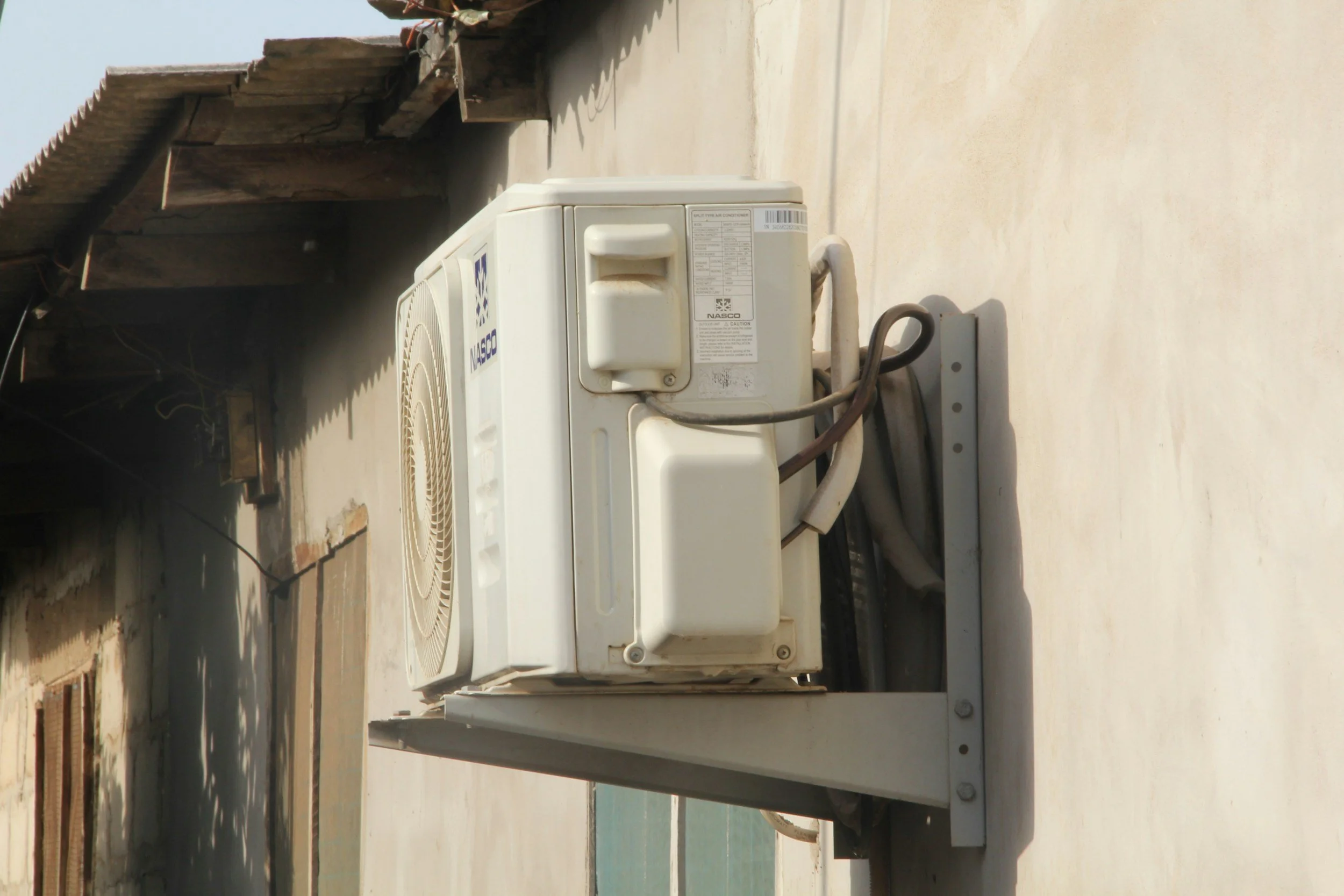 Image of an outdoor HVAC unit mounted on a building wall. The unit has a fan on top and labels, with electrical wiring connected to it. The background shows part of a roof and a sunny day.