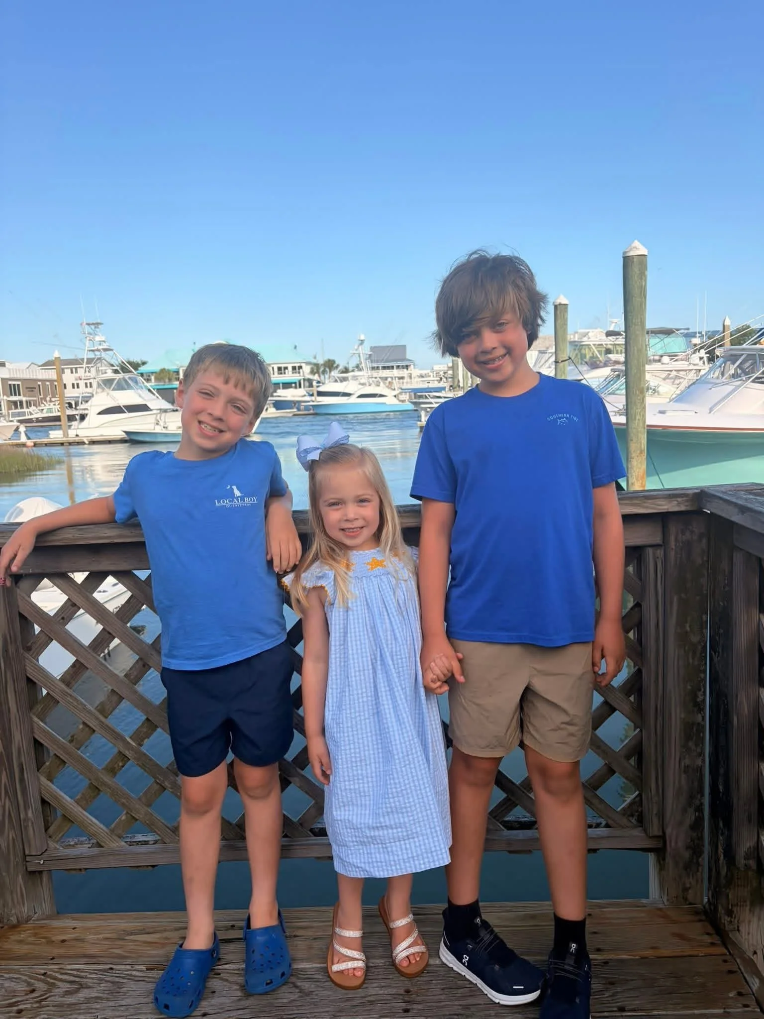 three children, two boys and a girl, standing on a wooden dock near boats in a harbor under a blue sky.