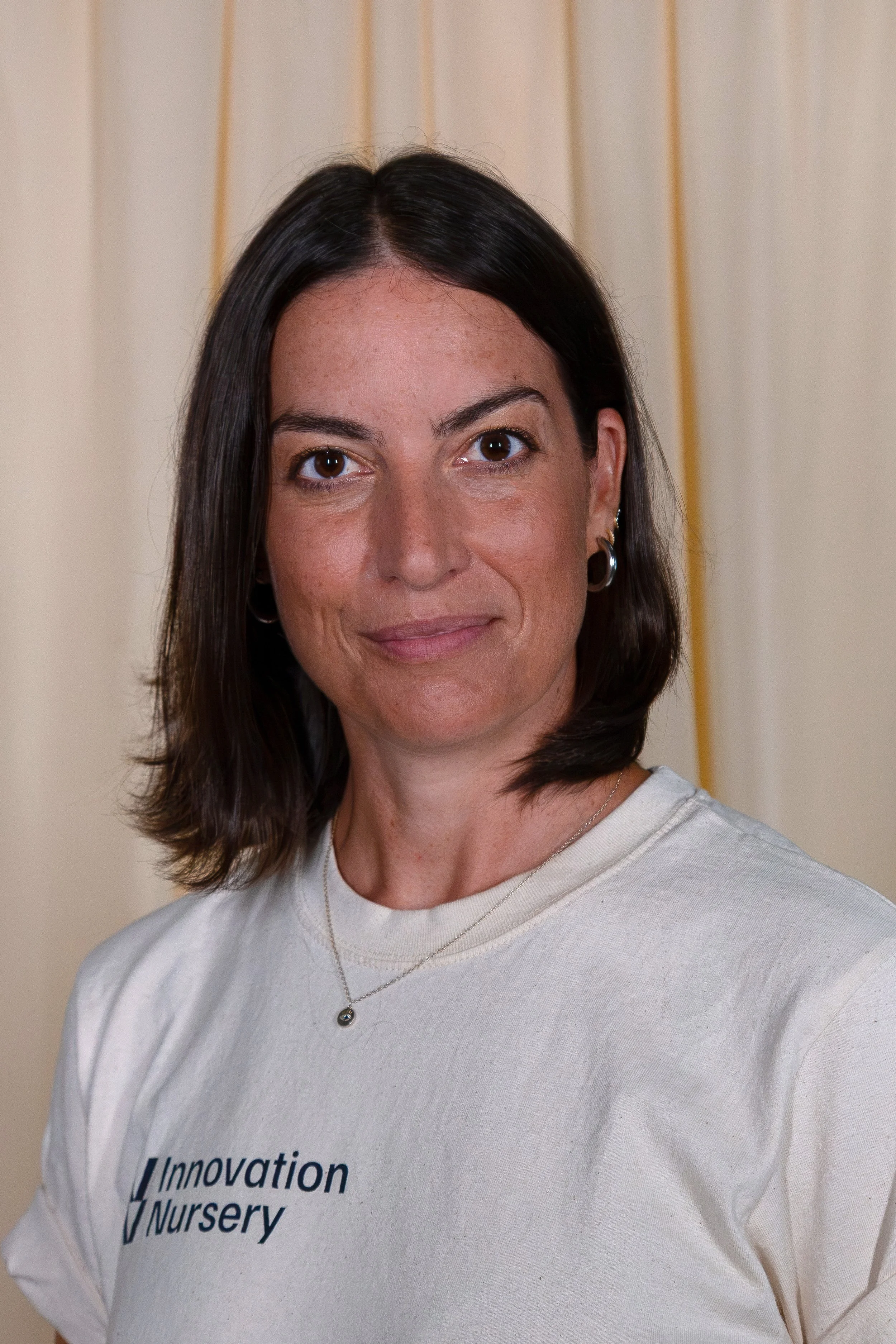 A woman with dark brown hair wearing a white shirt with partially visible text and a necklace with a small spherical pendant, standing against a beige background.